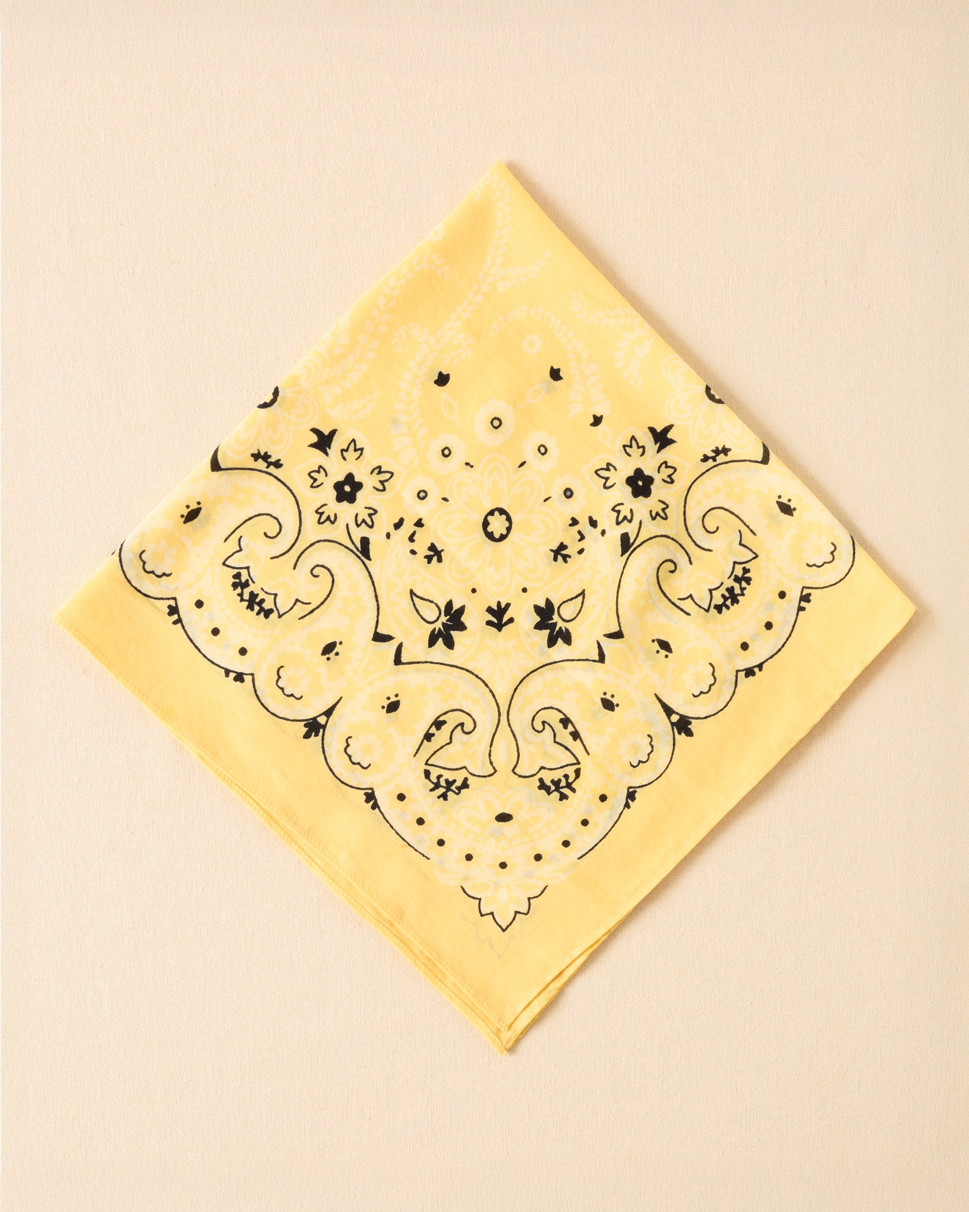 The Bodhi Sunlight Bandana, featuring a yellow base with a black and white paisley pattern, is neatly folded into a square and set against a plain beige background.