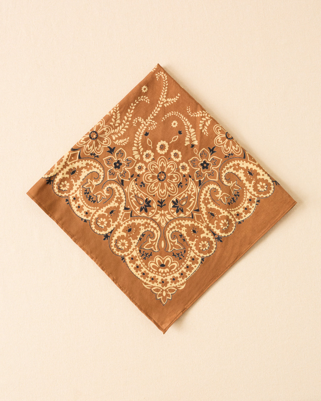 The Bodhi Toffee Bandana, a brown square cotton-silk accessory with cream and black floral paisley and botanical motifs, is shown laid flat on a light beige surface.