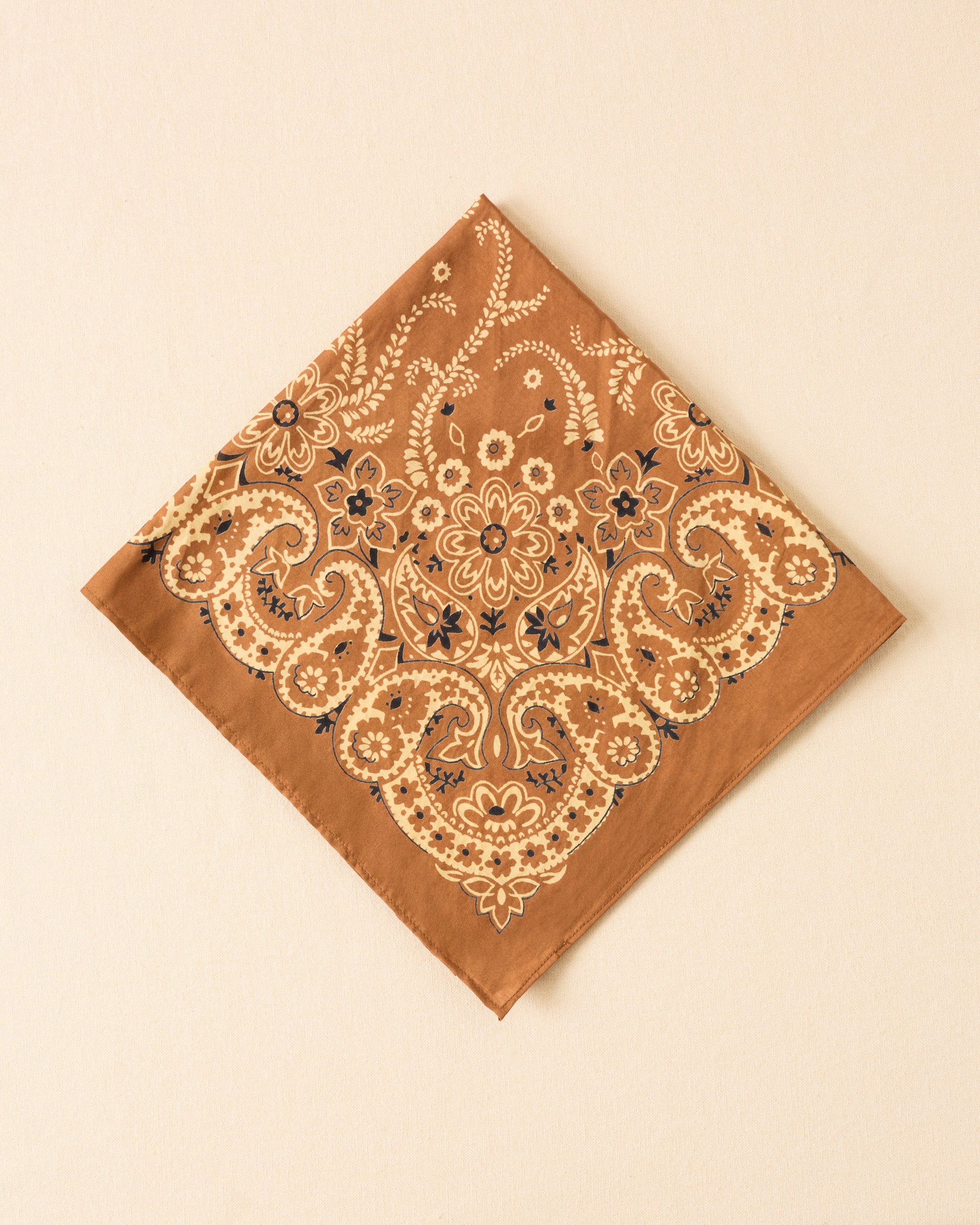 The Bodhi Toffee Bandana, a brown square cotton-silk accessory with cream and black floral paisley and botanical motifs, is shown laid flat on a light beige surface.