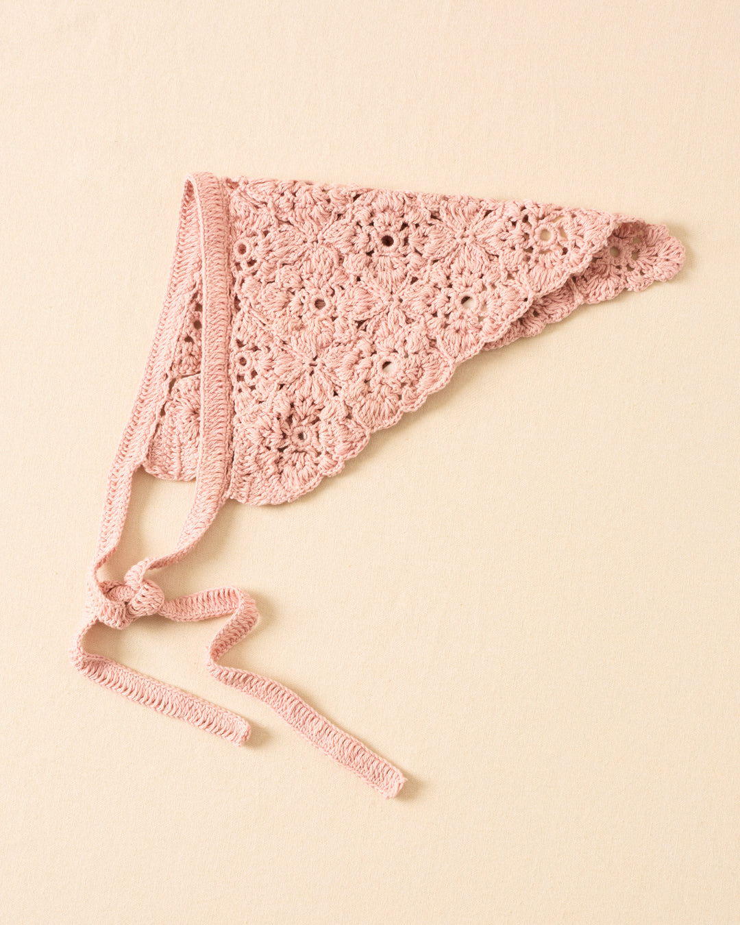The Bowie Ballet Crochet Bandana is a light pink cotton headscarf with a floral medallion motif, hand-crocheted with a delicate pattern and shown laying flat on a beige surface.