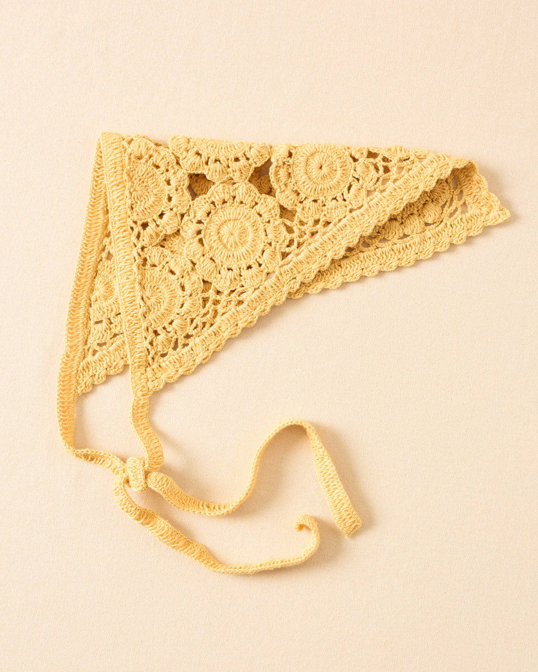 The Bowie Maize Crochet Bandana is a yellow cotton triangular headscarf with floral medallion patterns and two long ties, displayed flat on a beige surface.