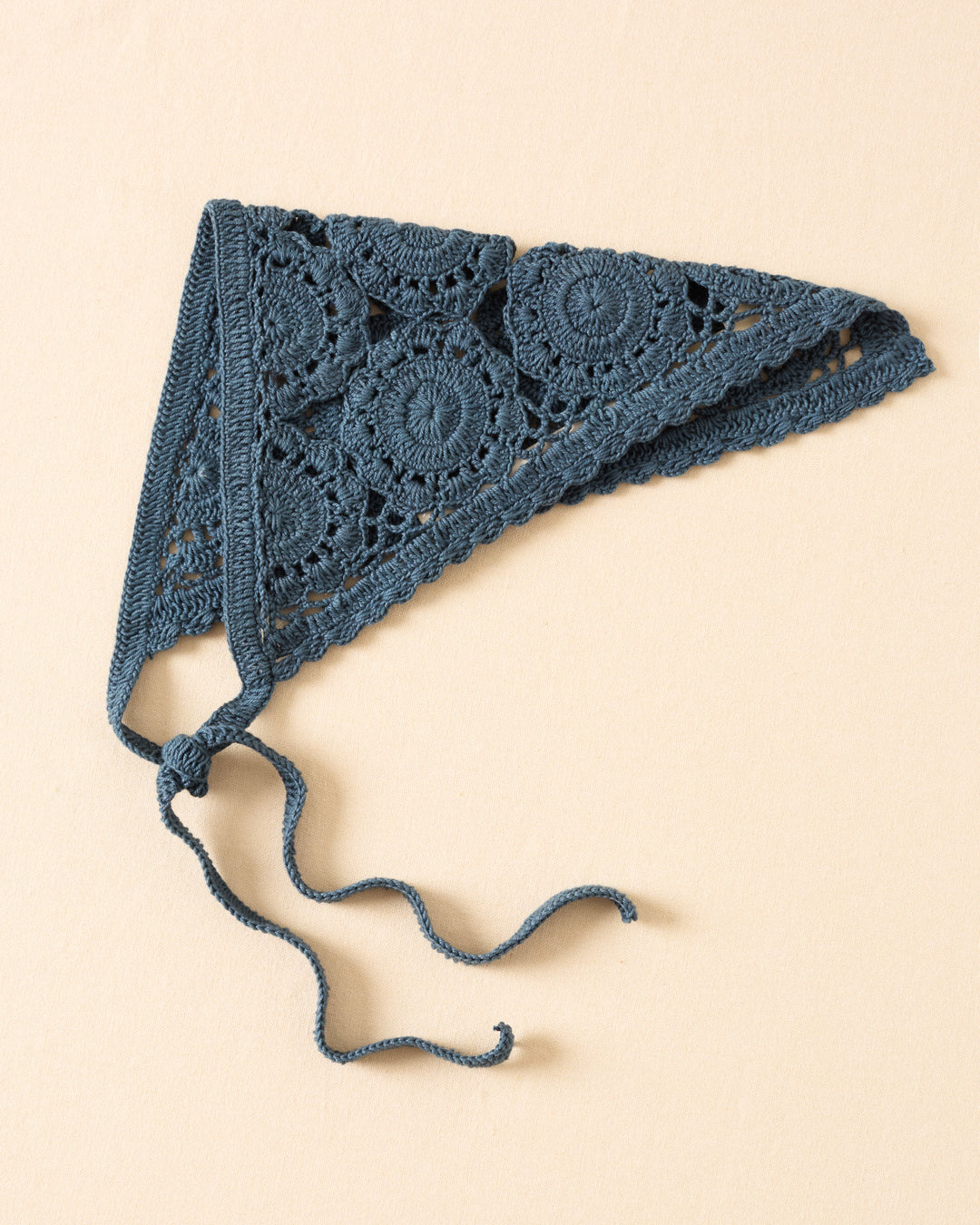 The Bowie Harbor Crochet Bandana is a blue, hand-crocheted triangle headscarf featuring circular floral medallions and two long ties, shown on a beige background.