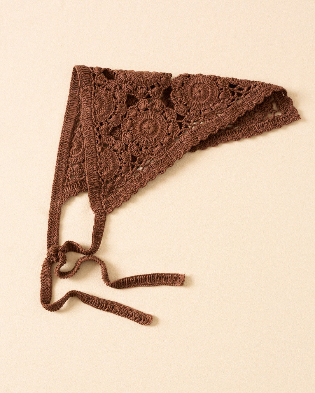 The Bowie Clove Crochet Bandana features intricate floral medallion patterns and two tie strings, displayed flat on a beige surface.