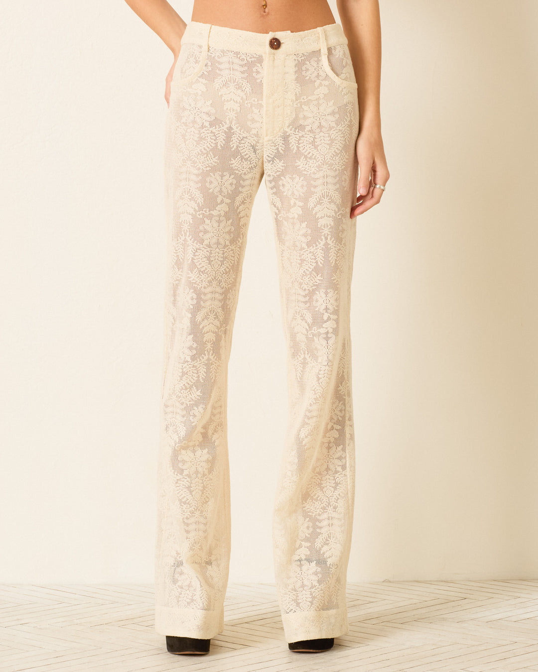 Someone models the Bunny Pearl Lace Lattice Jean, a high-waisted, semi-sheer cotton lace pant with a botanical motif and floral embroidery, and flared legs, standing in front of a plain light-colored wall.