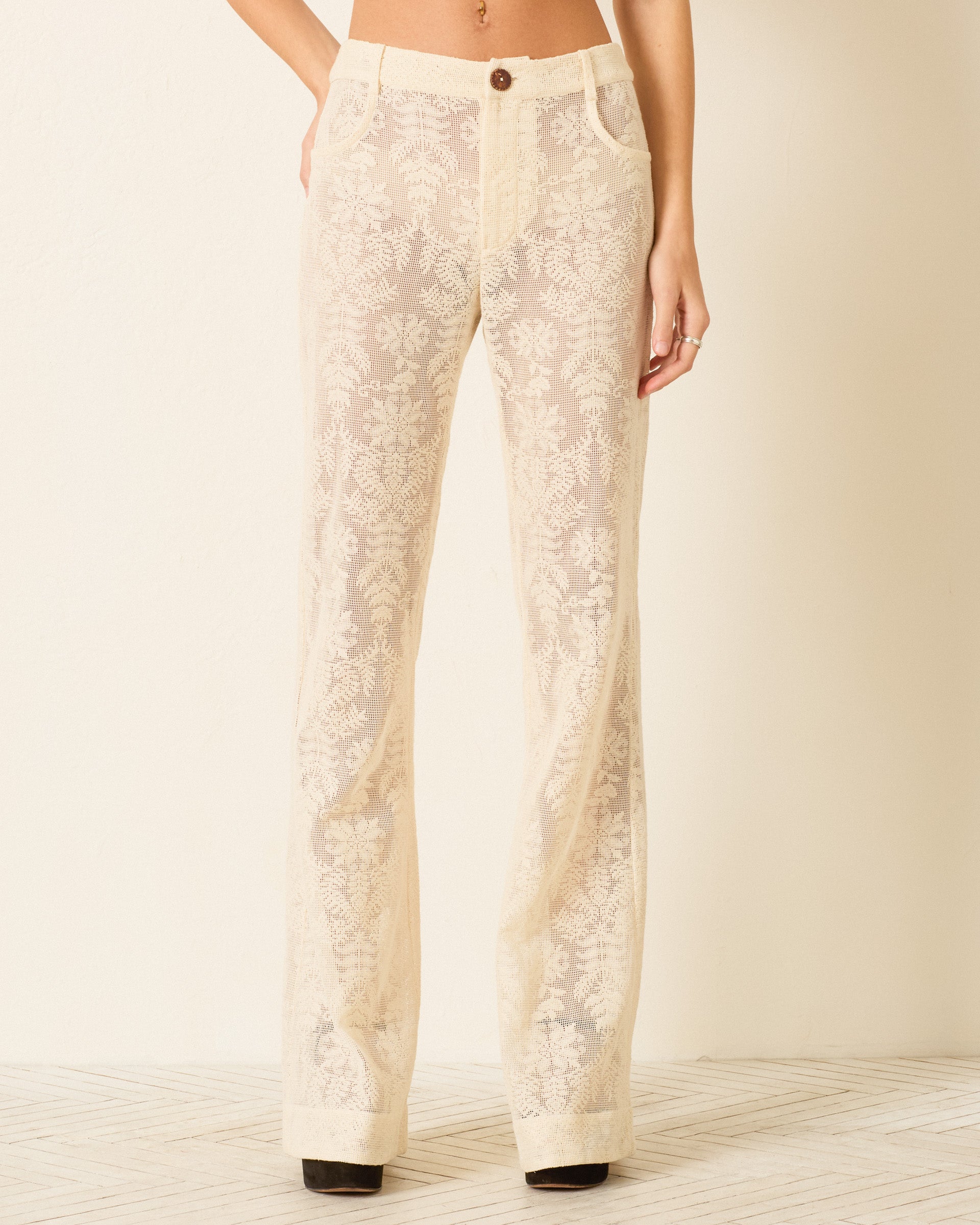 Someone models the Bunny Pearl Lace Lattice Jean, a high-waisted, semi-sheer cotton lace pant with a botanical motif and floral embroidery, and flared legs, standing in front of a plain light-colored wall.