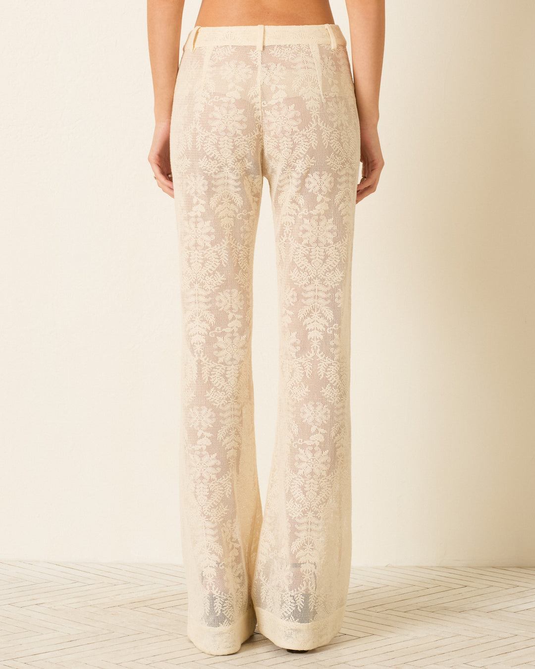 Person wearing the Bunny Pearl Lace Lattice Jean—a sheer, cream-colored cotton lace pant with a botanical motif and flared leg—viewed from the back, standing against a light-colored wall and floor.
