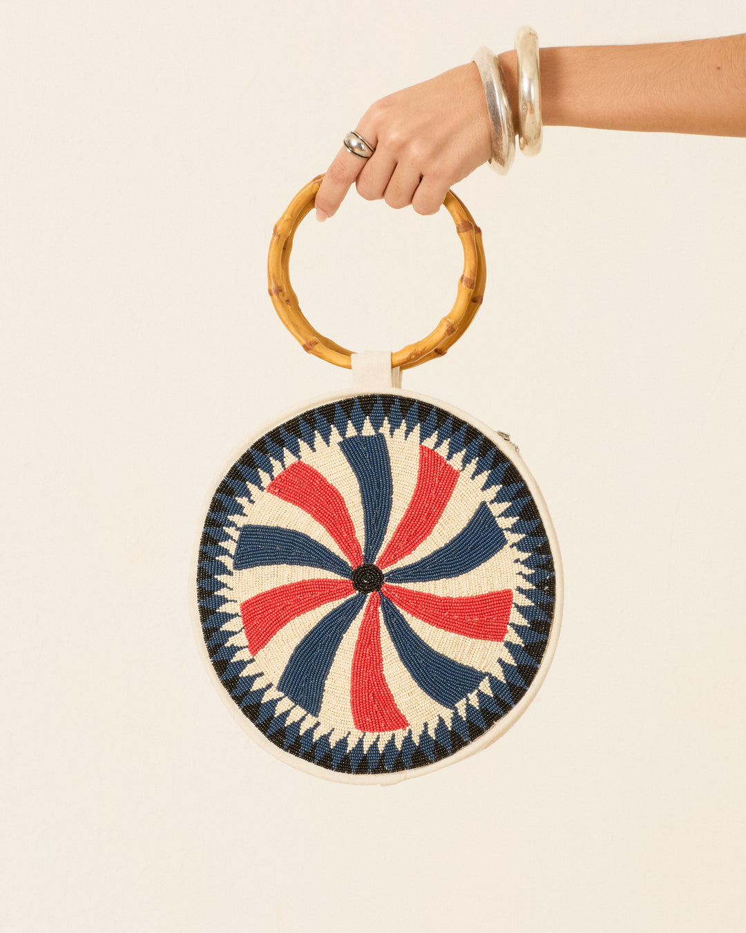 A hand holds the Carousel Candy Beaded Bag, featuring a bamboo handle and a blue, red, and white geometric spiral pattern on the front.
