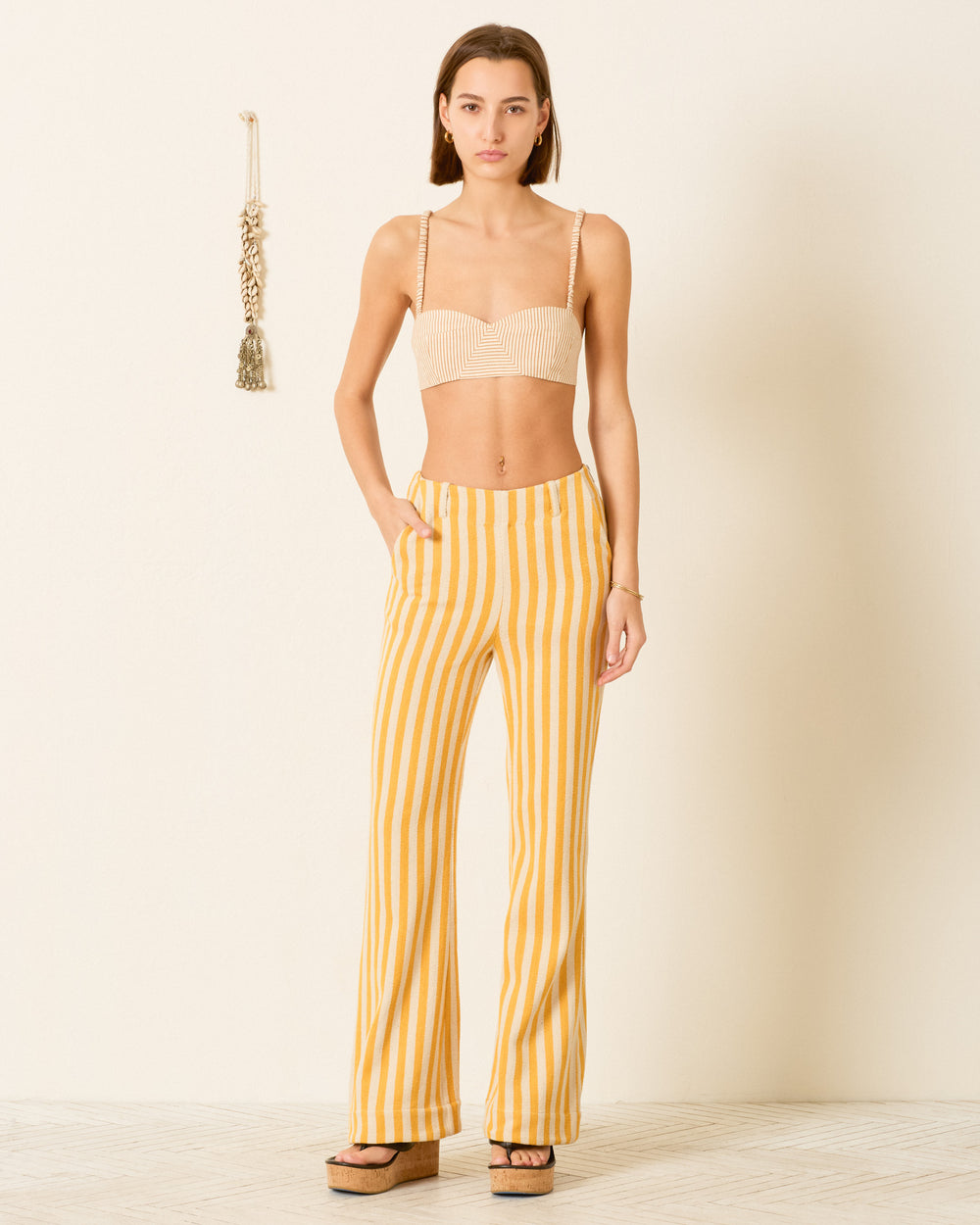 A woman stands against a plain wall wearing a beige crop top, the Charlie Sunlight Stripe Pant in yellow cotton twill, and platform sandals.