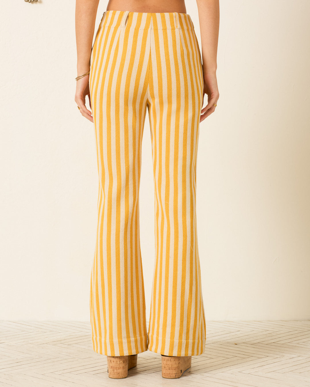 Model wears the Charlie Sunlight Stripe Pant—high-waisted, yellow striped cotton twill with flared legs—and platform sandals, seen from the back against a light background.