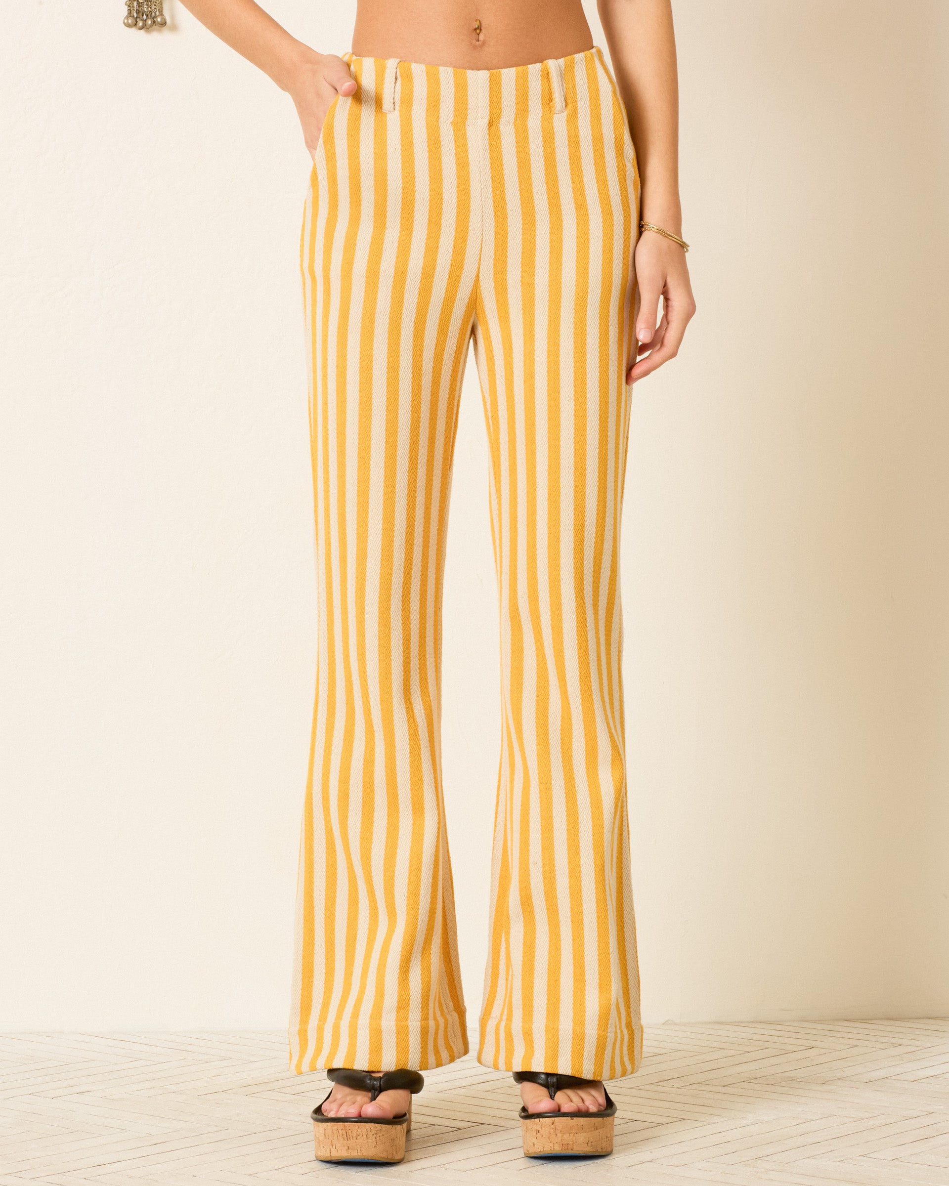 A person wearing the Charlie Sunlight Stripe Pant—high-waisted, yellow and white striped cotton twill flared pants—and black platform sandals, standing against a light background.