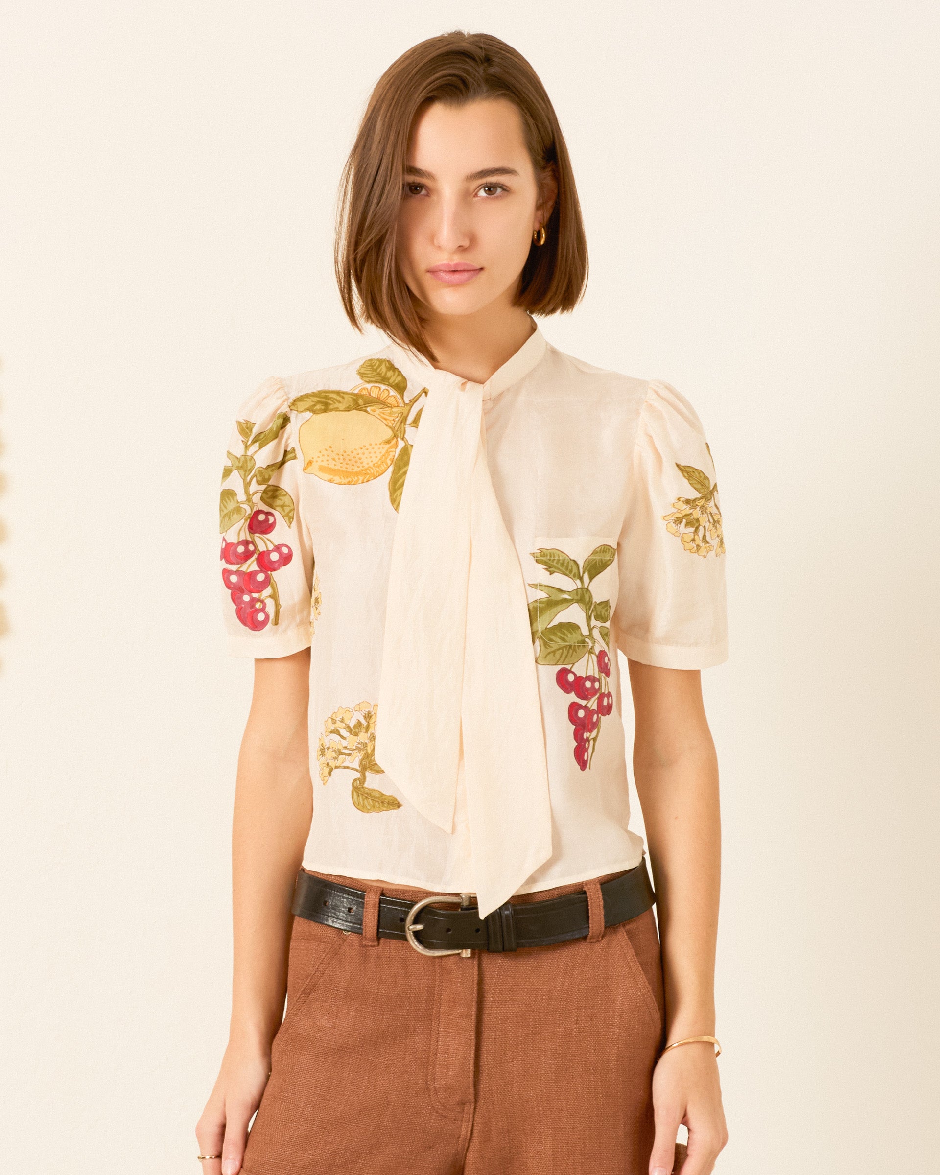 Wearing the Chance Four Fruit Silk Shirt—a cream, hand-loomed silk blouse featuring fruit and floral embroidery, puffed short sleeves, and a neck tie—paired with brown pants and a black belt, the person stands against a neutral background.