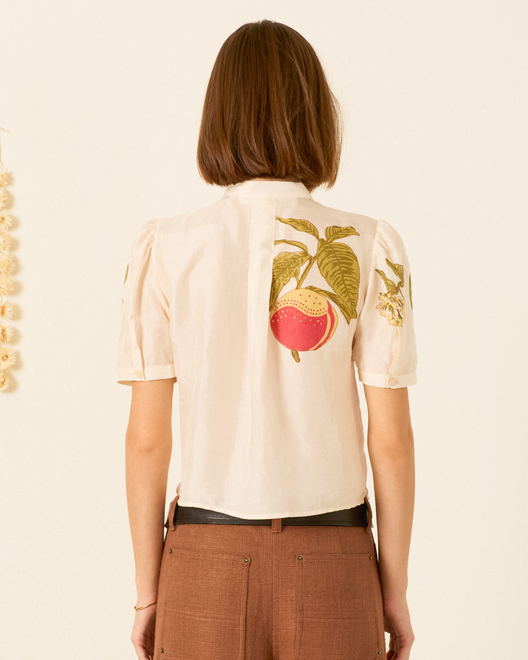 A person with short brown hair, wearing the Chance Four Fruit Silk Shirt—a light, short-sleeved blouse featuring block-printed fruit and leaf designs on the back—and brown pants, is shown from behind against a plain background.