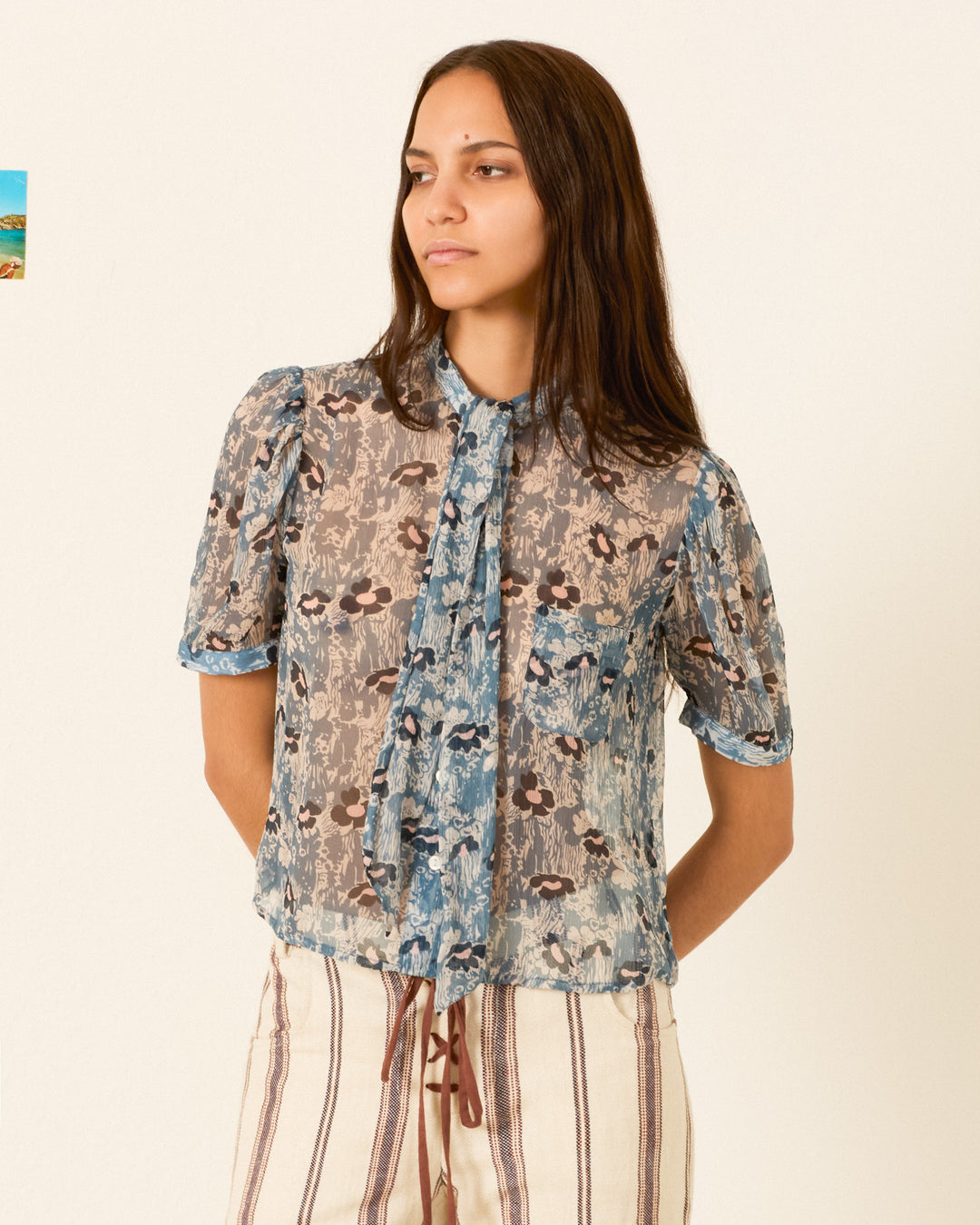 Wearing the Chance Water Flower Shirt, a sheer cupro-chiffon blouse adorned with a delicate floral motif and elegant pussy-bow ties at the neck, she pairs it with high-waisted, vertical striped pants and stands against a plain light background.