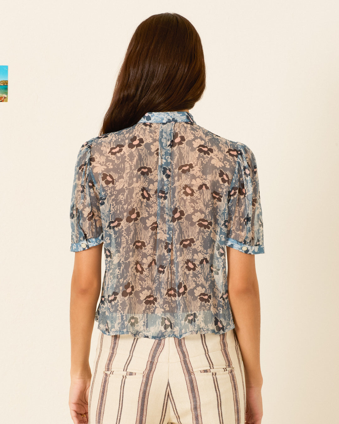 A woman stands with her back to the camera, wearing the Chance Water Flower Shirt—a sheer, short-sleeve cupro-chiffon blouse featuring a delicate floral motif—paired with high-waisted pants with vertical stripes.