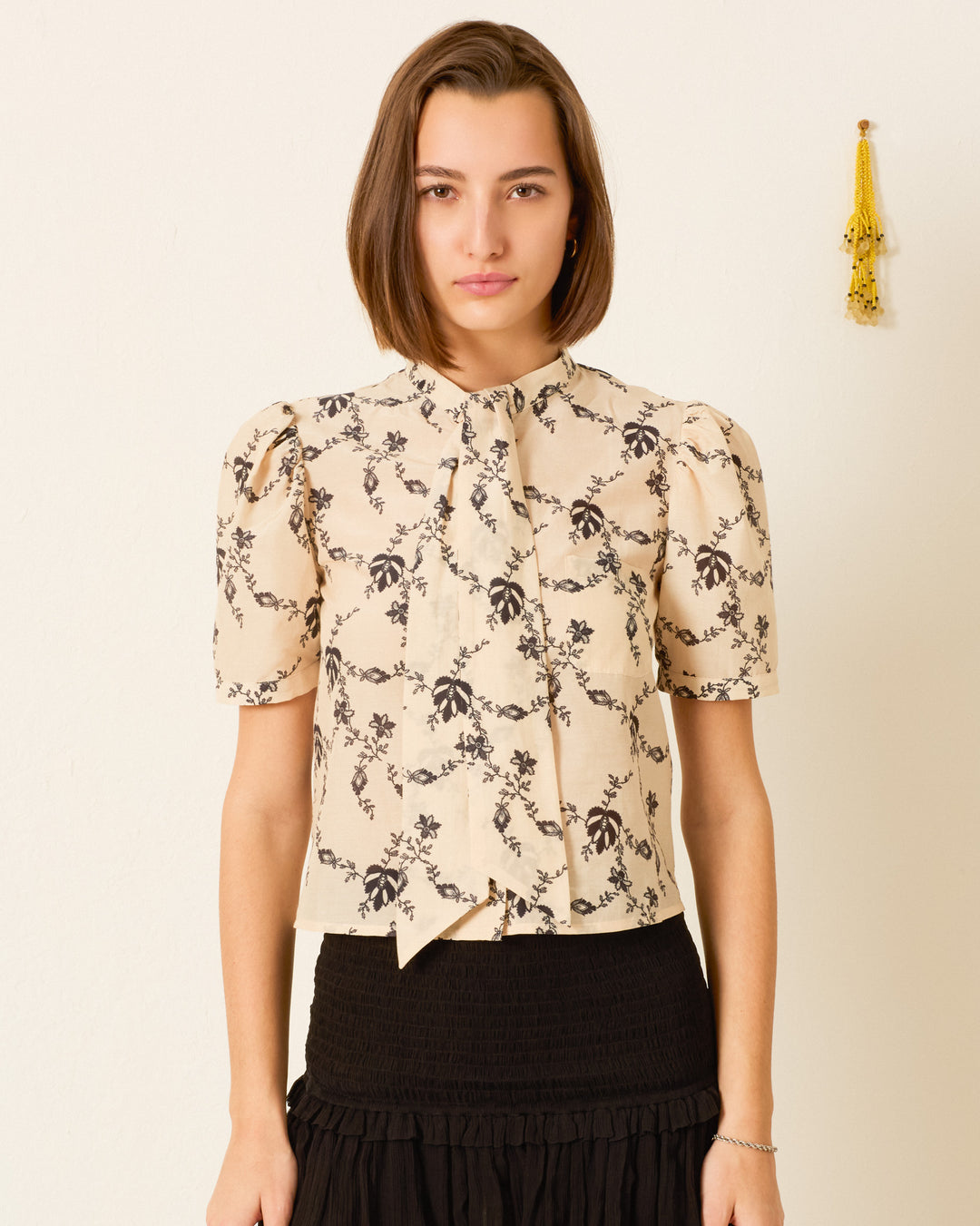 A woman wears a beige, short-sleeved Chance Shadow Lace Shirt with a black floral print, tucked into a black, tiered skirt.