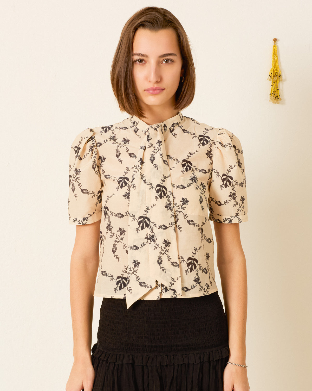 A woman wears a beige, short-sleeved Chance Shadow Lace Shirt with a black floral print, tucked into a black, tiered skirt.