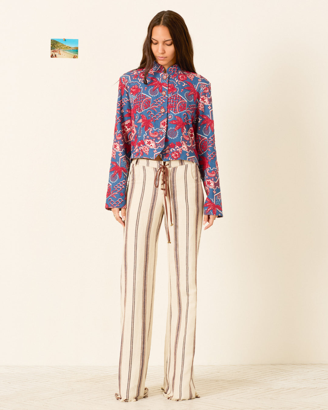 A woman stands against a plain background wearing the Clarke Delft Hive Jacket and high-waisted, wide-leg cream pants with vertical brown stripes.