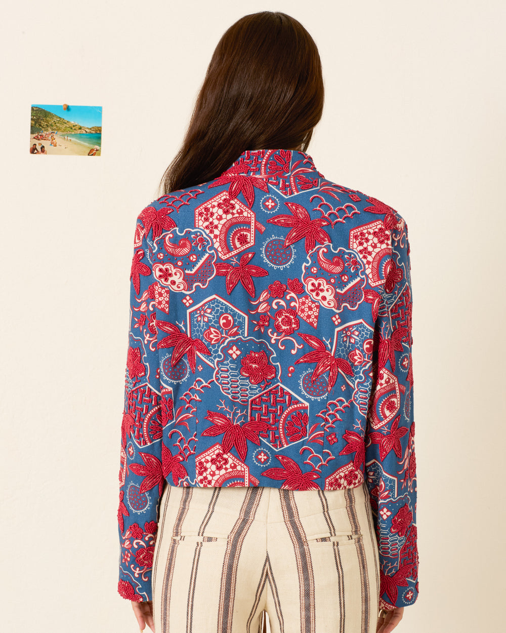 A person with long brown hair stands facing away, wearing the Clarke Delft Hive Jacket with blue and red wagara print, handmade floral buttons, and striped pants; a small beach photo hangs on the wall to the left.
