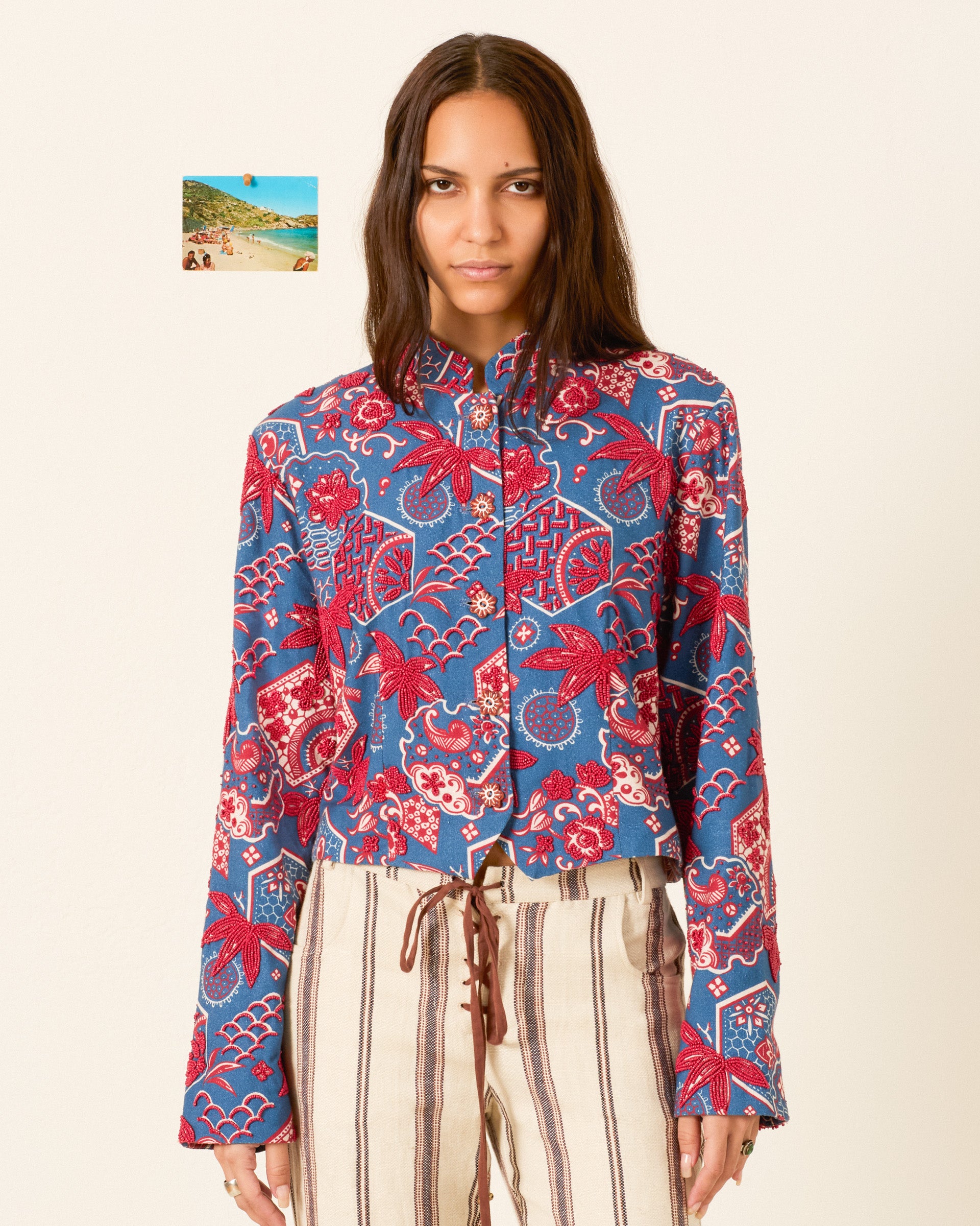 A woman stands against a plain background wearing the Clarke Delft Hive Jacket with a wagara print and handmade floral buttons, paired with beige striped pants; a small beach photo hangs on the wall behind her.