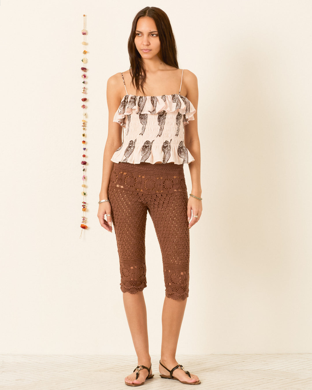 A woman stands indoors wearing the Claire Snail Study Silk Top, a hand-block-printed silk garment featuring a bird print and a ruffled peplum hem. She pairs it with cropped brown crochet pants and sandals. Decorative hanging beads are visible in the background.