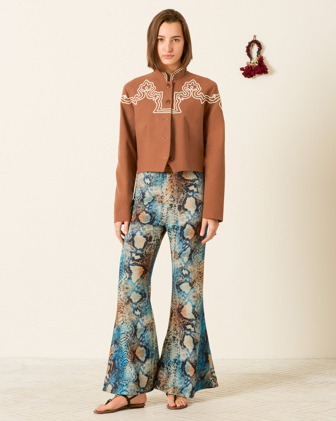 A woman stands against a plain wall wearing the Clarke Clove Spur Jacket, featuring white embroidery and silver studs, along with flared blue patterned pants and sandals. A small decorative wreath hangs on the wall behind her.