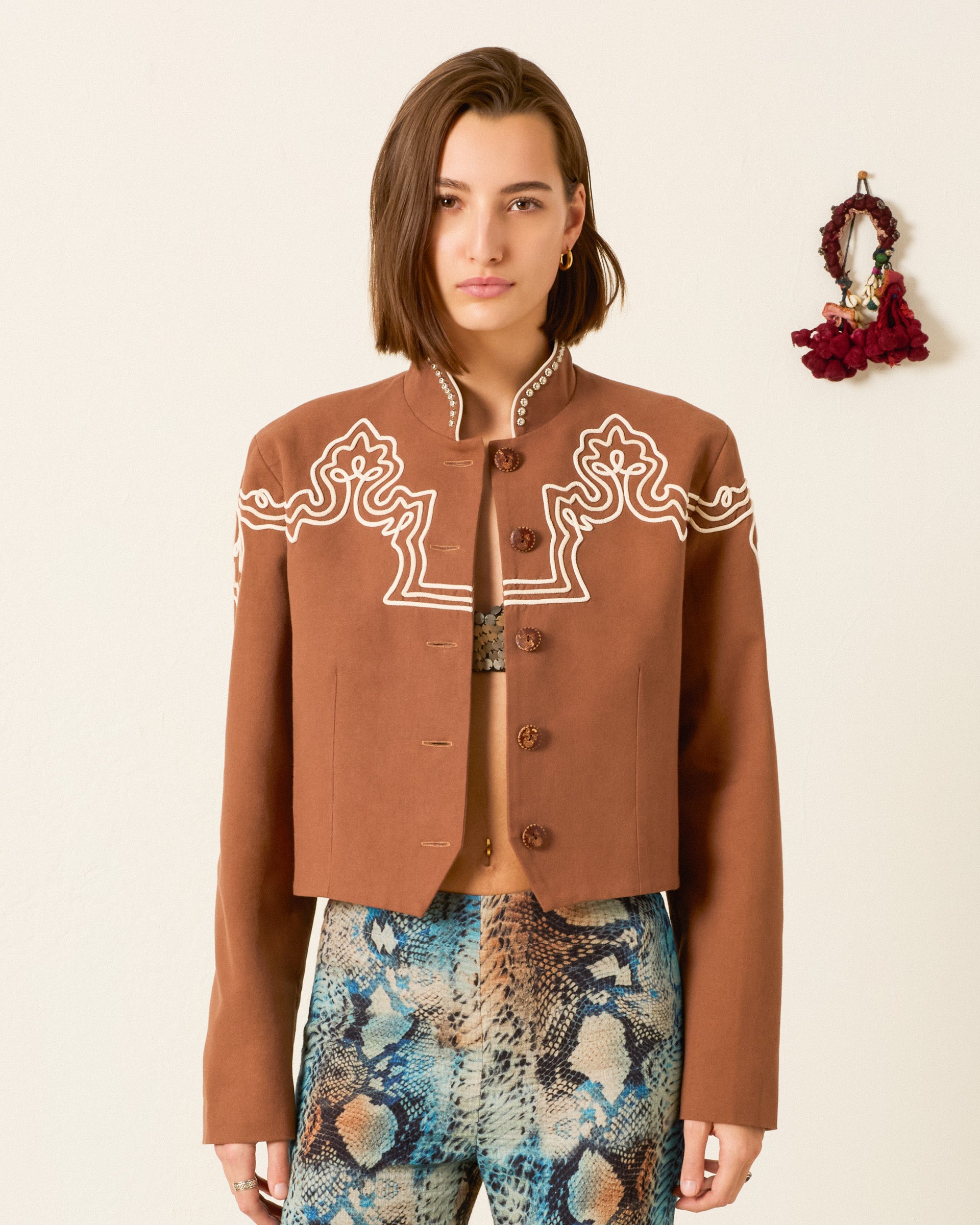 A woman faces forward wearing the Clarke Clove Spur Jacket—a brown cropped jacket with white embroidery and silver studs—paired with patterned pants, in front of a plain light wall decorated with a small hanging item.