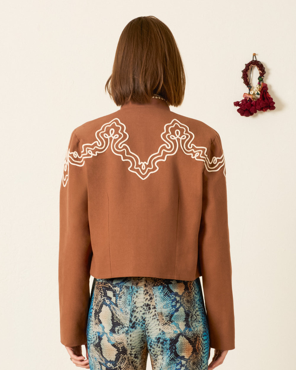 A person stands facing away, wearing the Clarke Clove Spur Jacket—a cropped brown jacket with white trim and silver studs—paired with multicolored snakeskin-patterned pants. A small wall decoration hangs to the right.