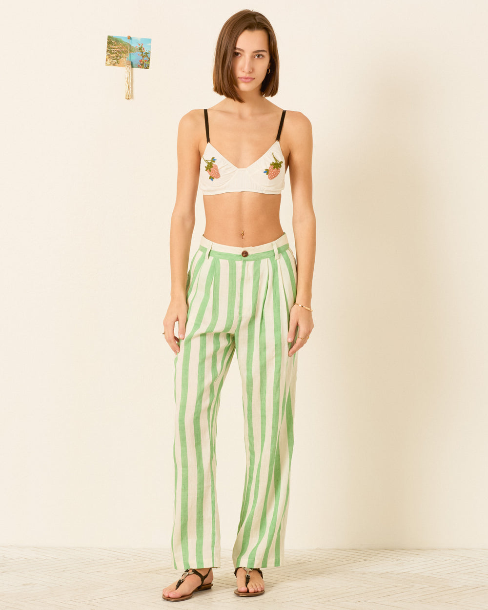 A woman stands indoors wearing a white bralette with floral embroidery, the Colette Celery Stripe Pant in a relaxed silhouette, and sandals. A small photo is pinned to the wall behind her.