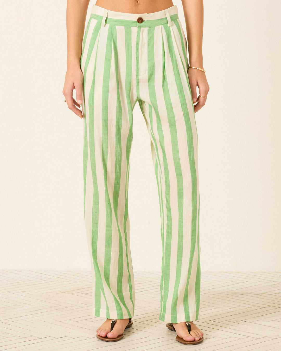 A person is wearing the Colette Celery Stripe Pant, a high-waisted, wide-leg style featuring vertical green and white stripes in a linen-silk blend. Shown from waist to feet while standing on a light patterned floor, the relaxed silhouette of these pants adds effortless sophistication to the overall look.
