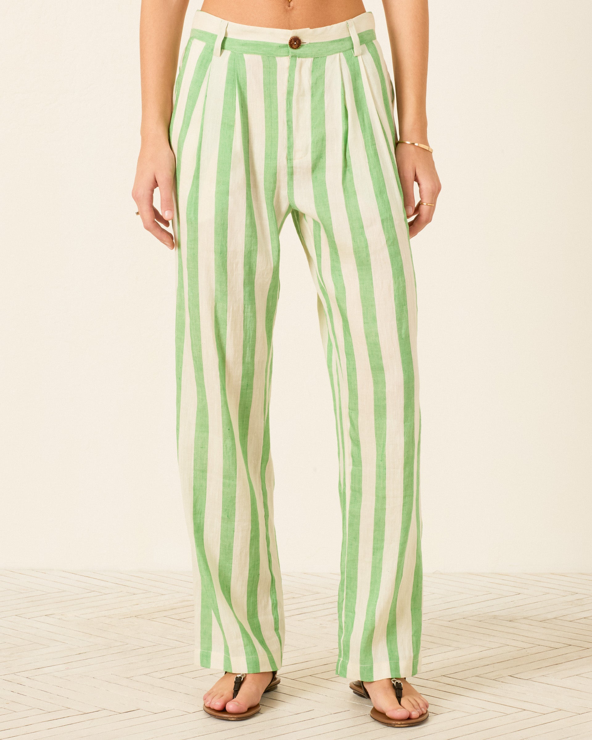 A person is wearing the Colette Celery Stripe Pant, a high-waisted, wide-leg style featuring vertical green and white stripes in a linen-silk blend. Shown from waist to feet while standing on a light patterned floor, the relaxed silhouette of these pants adds effortless sophistication to the overall look.