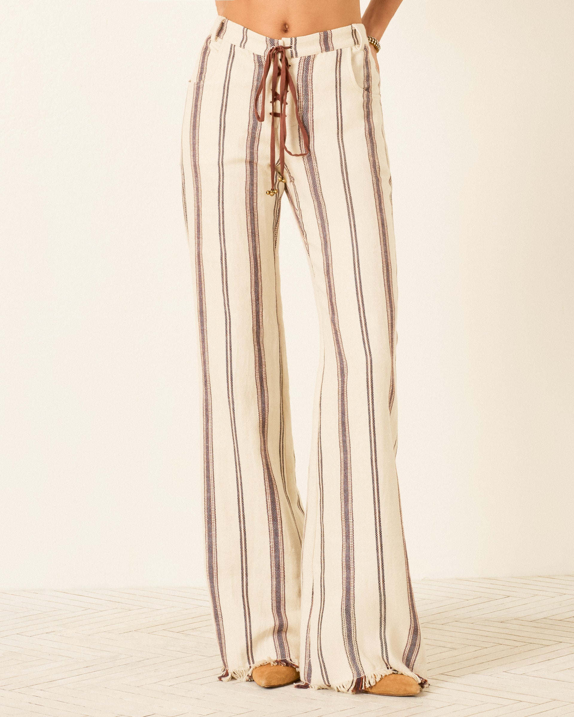 Wearing the Cooper Bark Stripe Pant—cream-colored, flared linen-cotton pants with brown and gray vertical stripes, front tie closure, and raw hem—standing on a light wood floor.