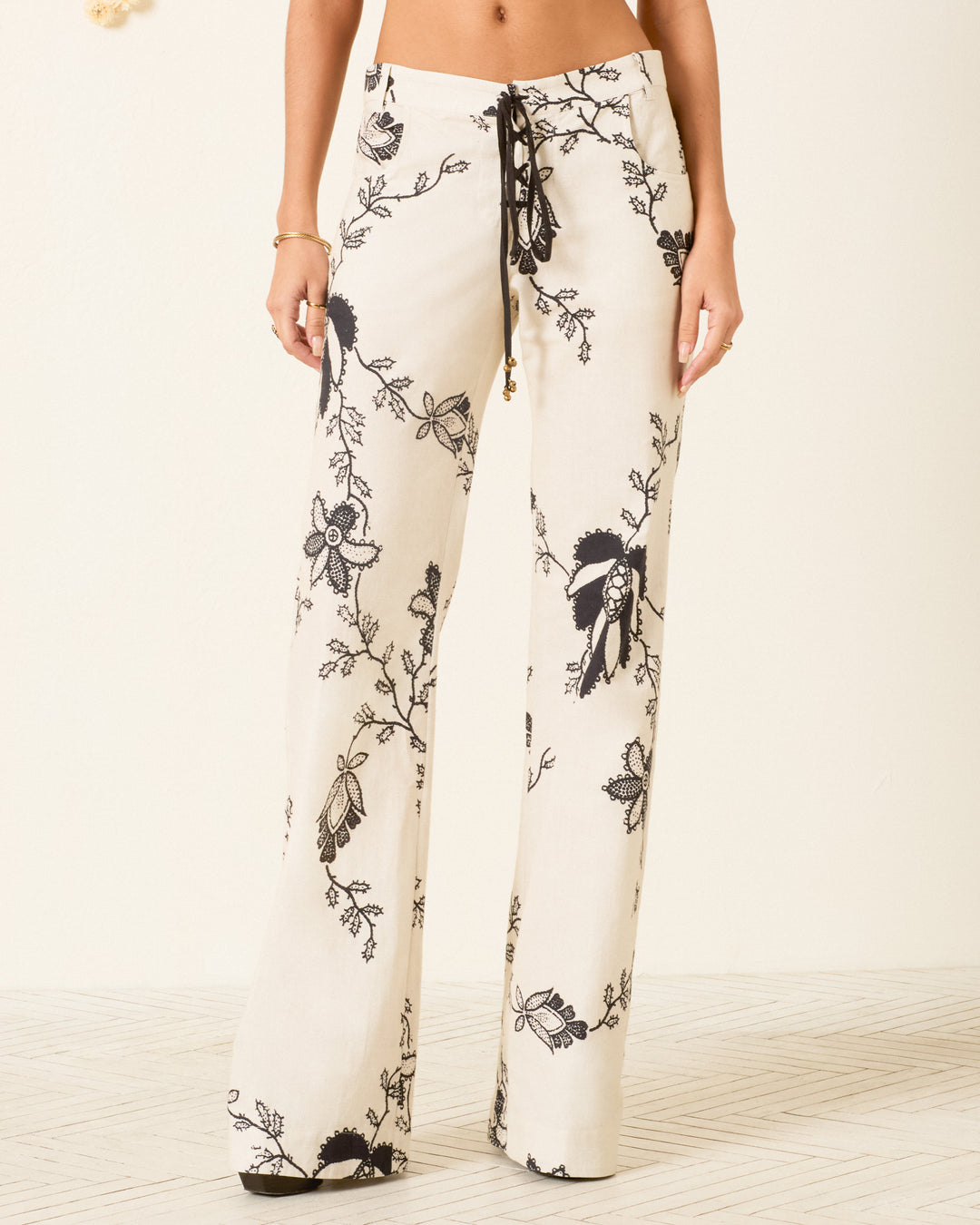A person wearing the Cooper Shadow Lace Pant—high-waisted, wide-leg cream khadi cotton pants with a black floral print and drawstring waist—stands on a light-patterned floor.