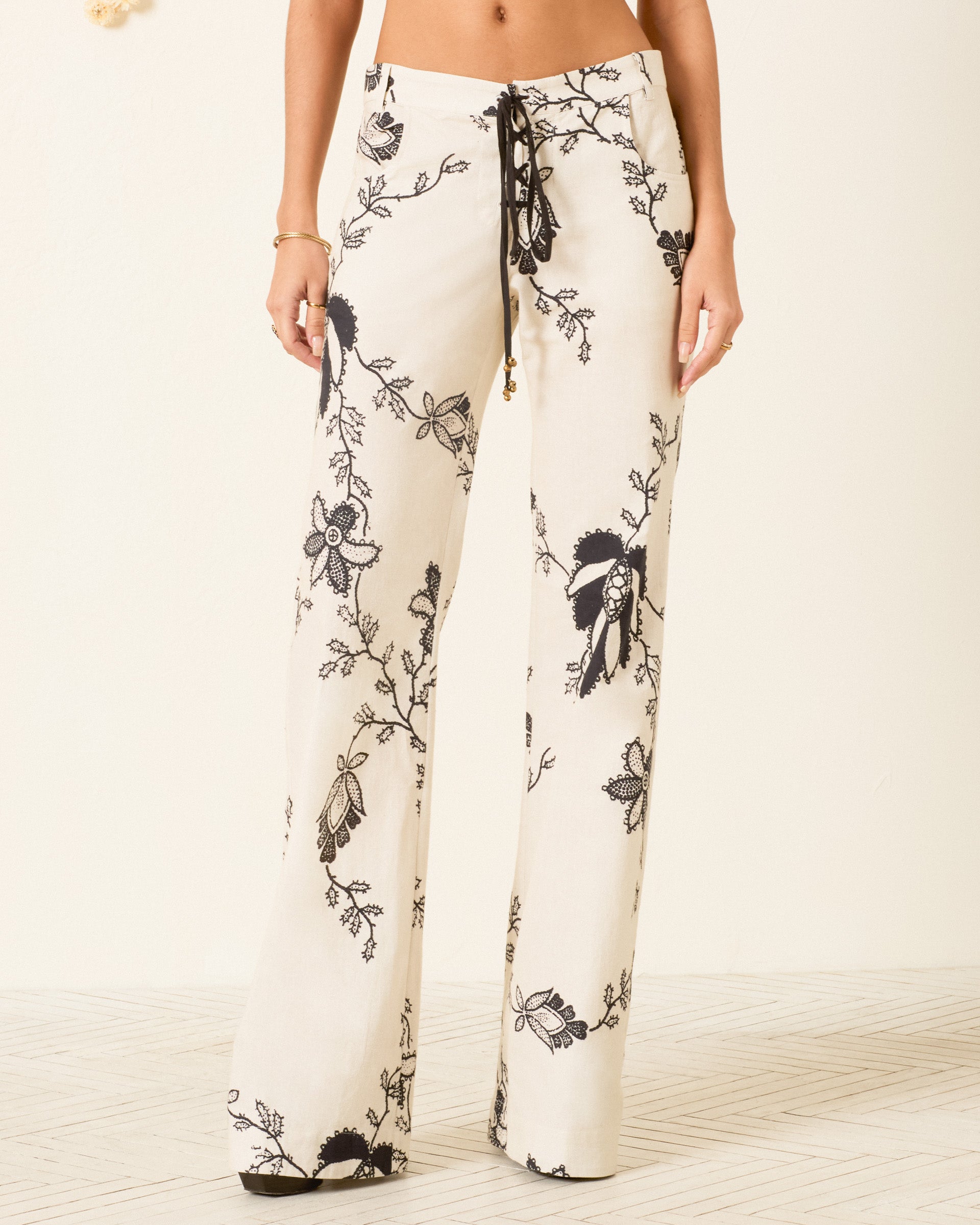A person wearing the Cooper Shadow Lace Pant—high-waisted, wide-leg cream khadi cotton pants with a black floral print and drawstring waist—stands on a light-patterned floor.