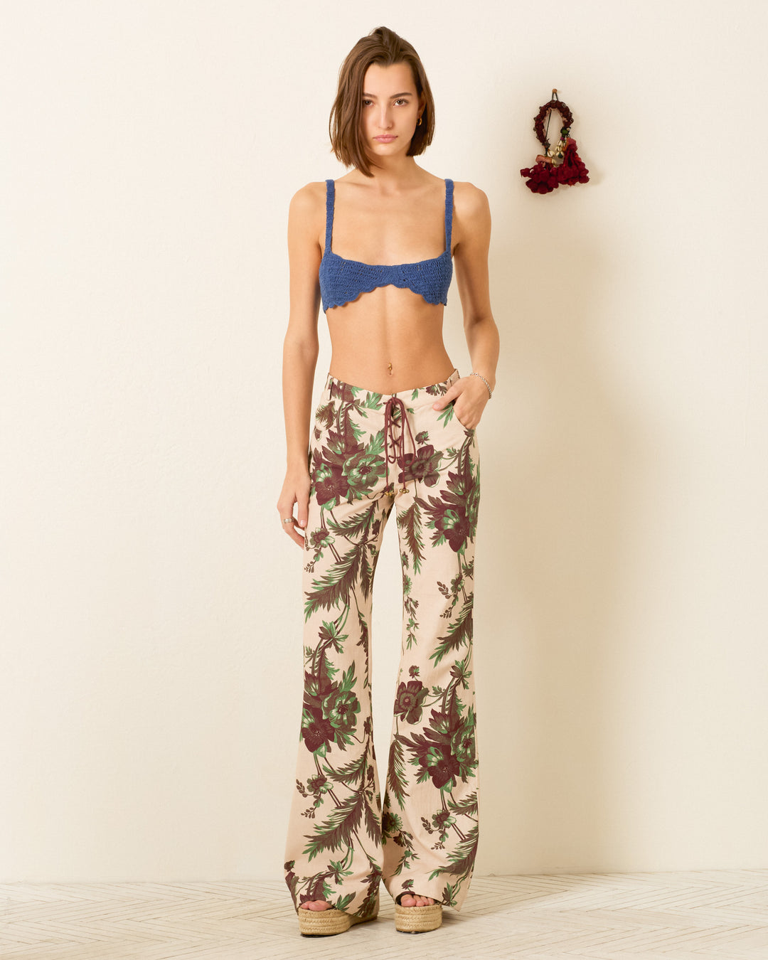 A woman stands indoors wearing the Cooper Summermoss Pant, a high-waisted lace-up style with a botanical motif, paired with a blue bralette and wedge sandals. A small decorative wreath decorates the wall behind her.
