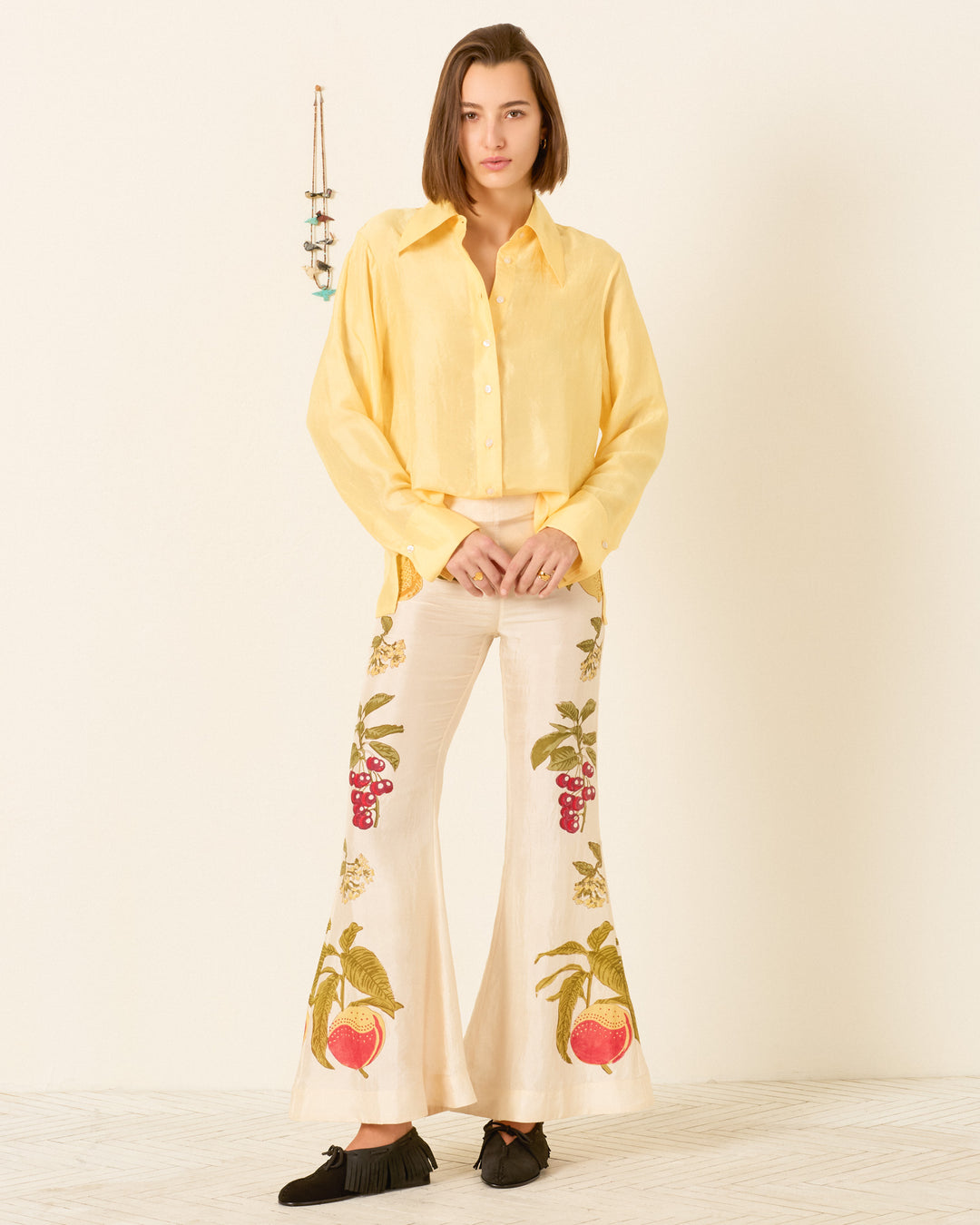 A person stands indoors wearing the Dana Sunlight Silk Shirt, a vintage-inspired yellow button-up with a 70s dagger collar, paired with cream flared pants featuring fruit and floral embroidery and black shoes.
