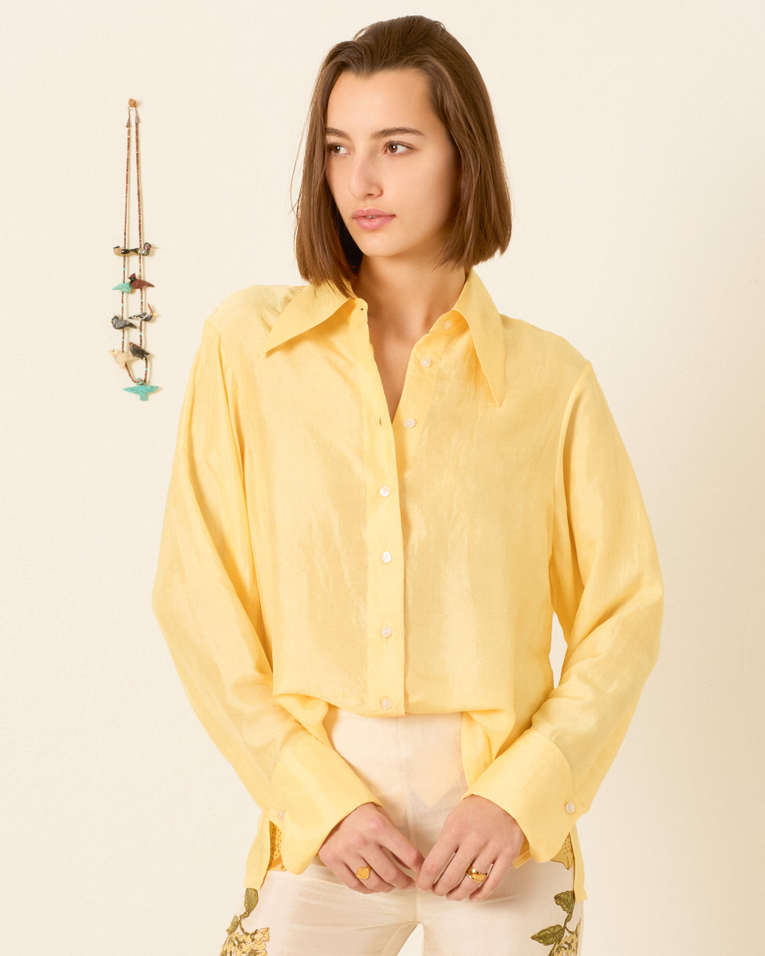 A woman with straight brown hair wears the Dana Sunlight Silk Shirt—a vintage-inspired yellow button-up with a ’70s dagger collar—paired with light pants, standing against a plain wall with a necklace hanging on the left.