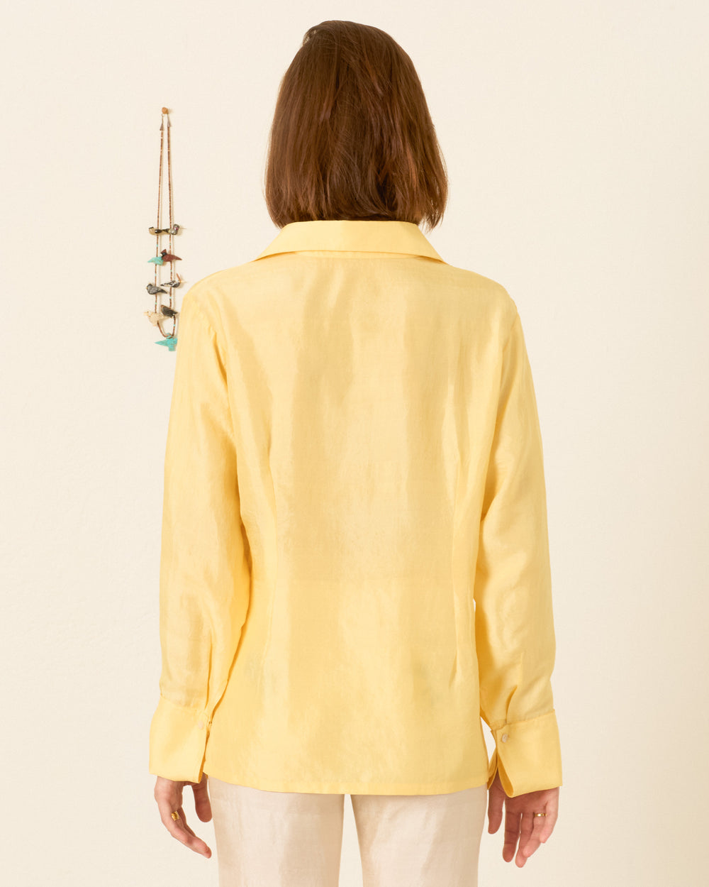 A person with shoulder-length brown hair, wearing the Dana Sunlight Silk Shirt and beige pants, stands facing away from the camera. A small hanging decoration is on the wall.