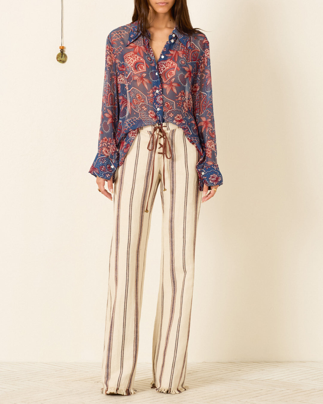 A person wearing the Dana Delft Hive Shirt, a blue and red wagara-print button-up, paired with beige wide-leg pants with vertical brown stripes, stands against a plain light-colored wall.