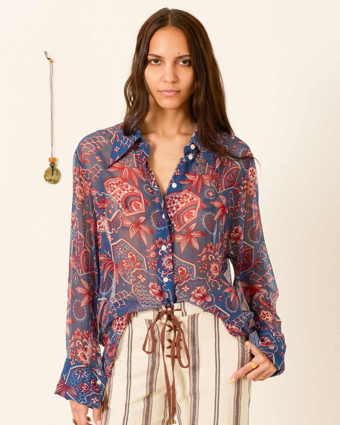 A woman wears the Dana Delft Hive Shirt, a sheer cupro-chiffon, long-sleeve blouse with floral patterns and a 70s dagger collar, paired with high-waisted striped pants, standing against a plain wall with a small hanging ornament.