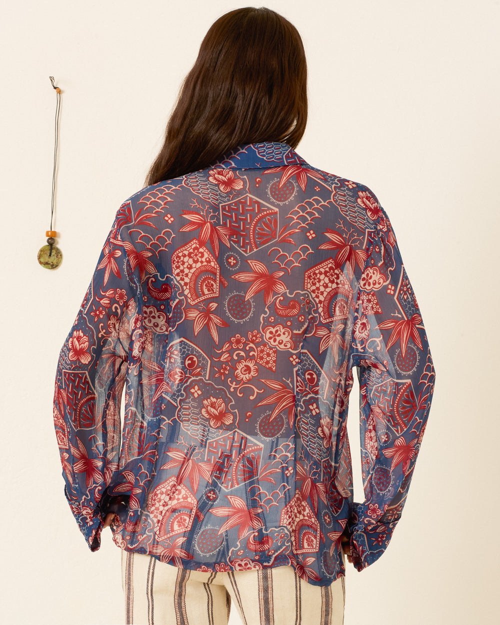A person stands with their back to the camera, wearing the Dana Delft Hive Shirt—sheer cupro-chiffon with a red and blue floral wagara print—and striped pants. A small decorative item hangs on the wall.