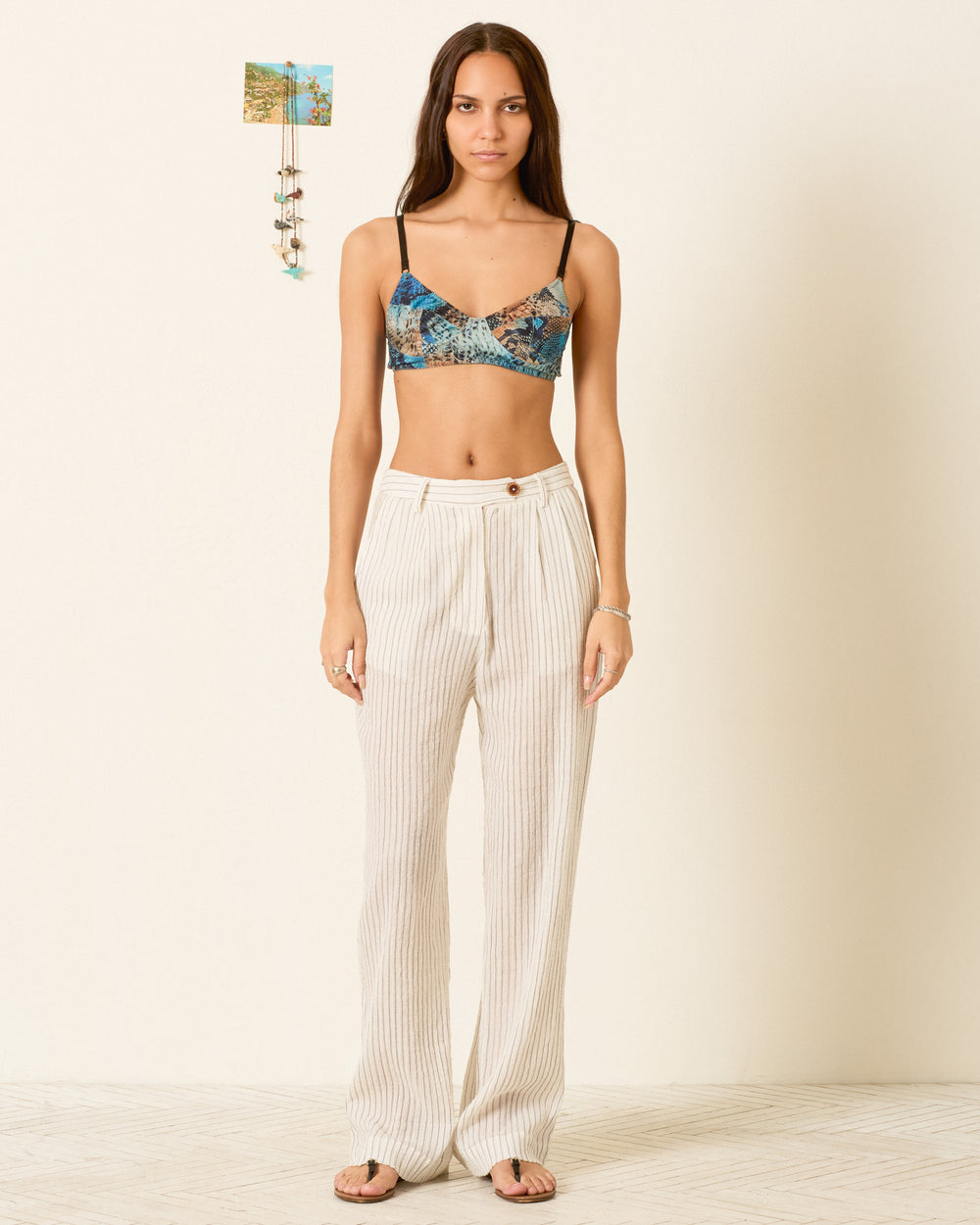 A woman stands indoors wearing a patterned crop top, Diana Sea Salt Pant in a relaxed fit gauzy cotton stripe, and sandals, with a small photo artwork displayed on the wall behind her.