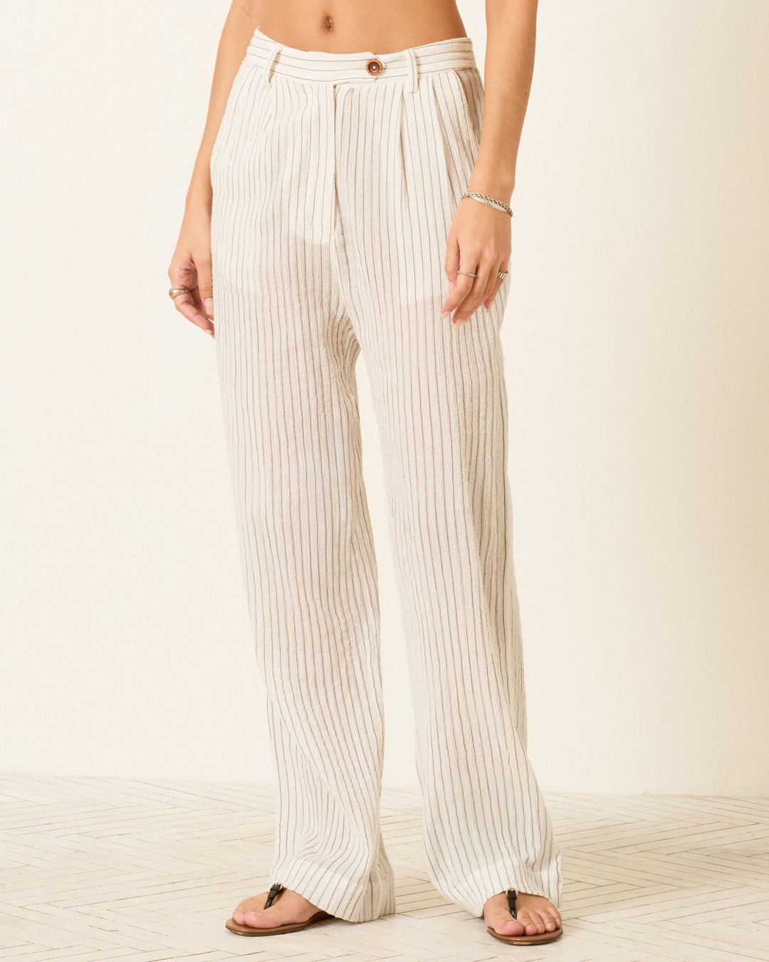 A person wearing Diana Sea Salt Pant—high-waisted, white, wide-leg pants with yarn-dyed stripes, button closure, and a relaxed fit—stands barefoot on a light-colored floor.