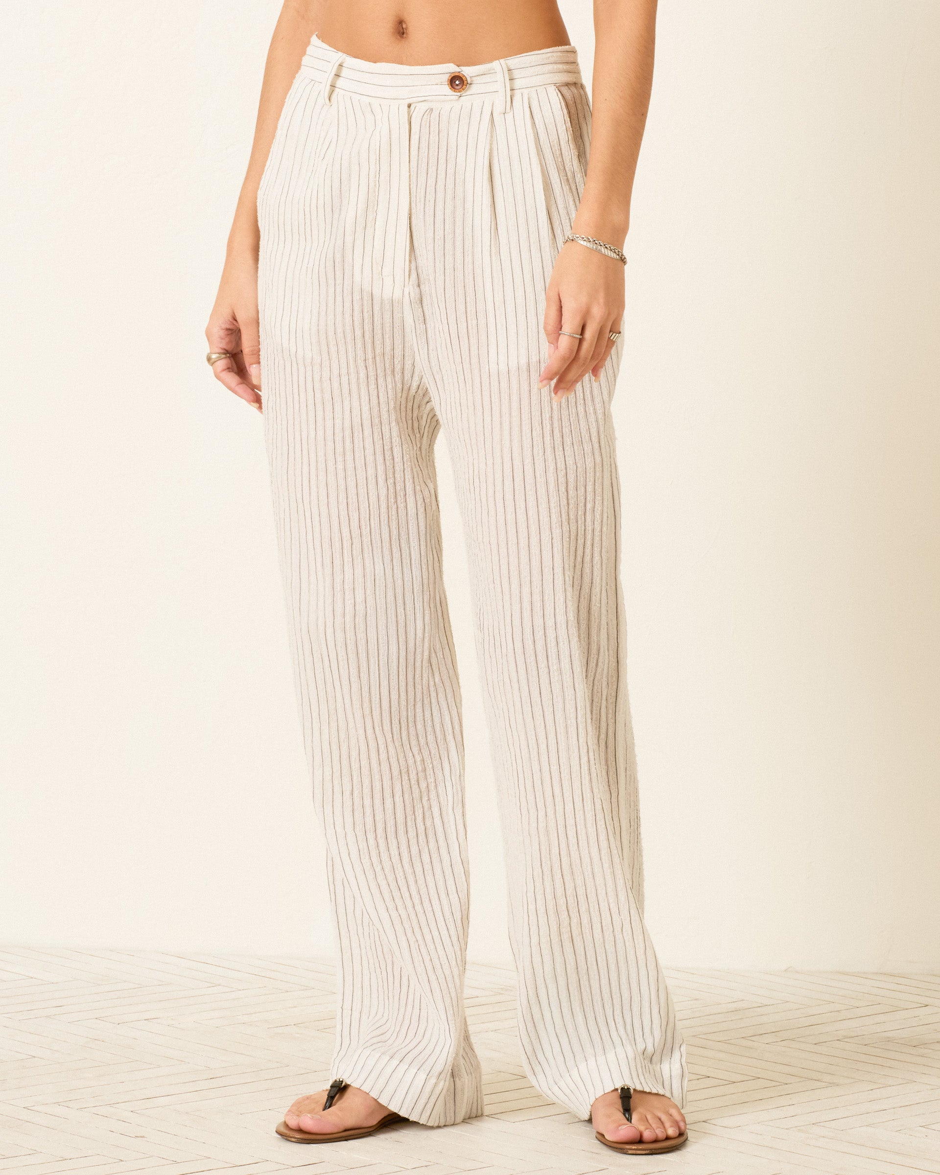 A person wearing Diana Sea Salt Pant—high-waisted, white, wide-leg pants with yarn-dyed stripes, button closure, and a relaxed fit—stands barefoot on a light-colored floor.