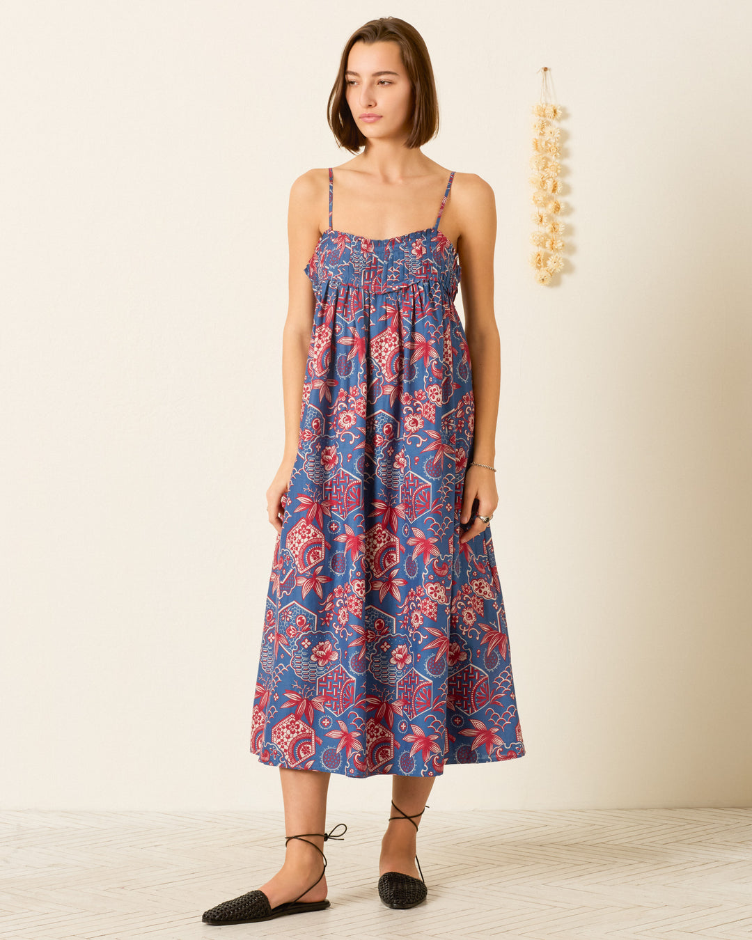 A woman stands indoors wearing the Eden Delft Hive Dress, a blue and red wagara print sleeveless maxi with thin straps, paired with black flats. Dried flowers hang on the wall behind her.