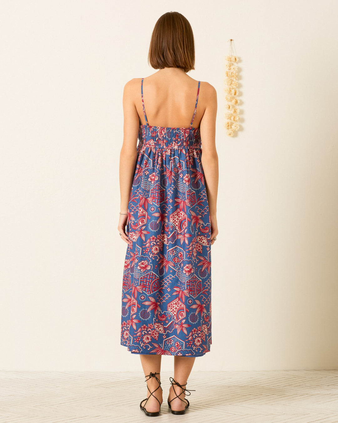 A woman stands indoors with her back to the camera, wearing the Eden Delft Hive Dress—a blue and red cotton-silk blend maxi sundress with thin straps—and black sandals. A wall decoration hangs nearby.