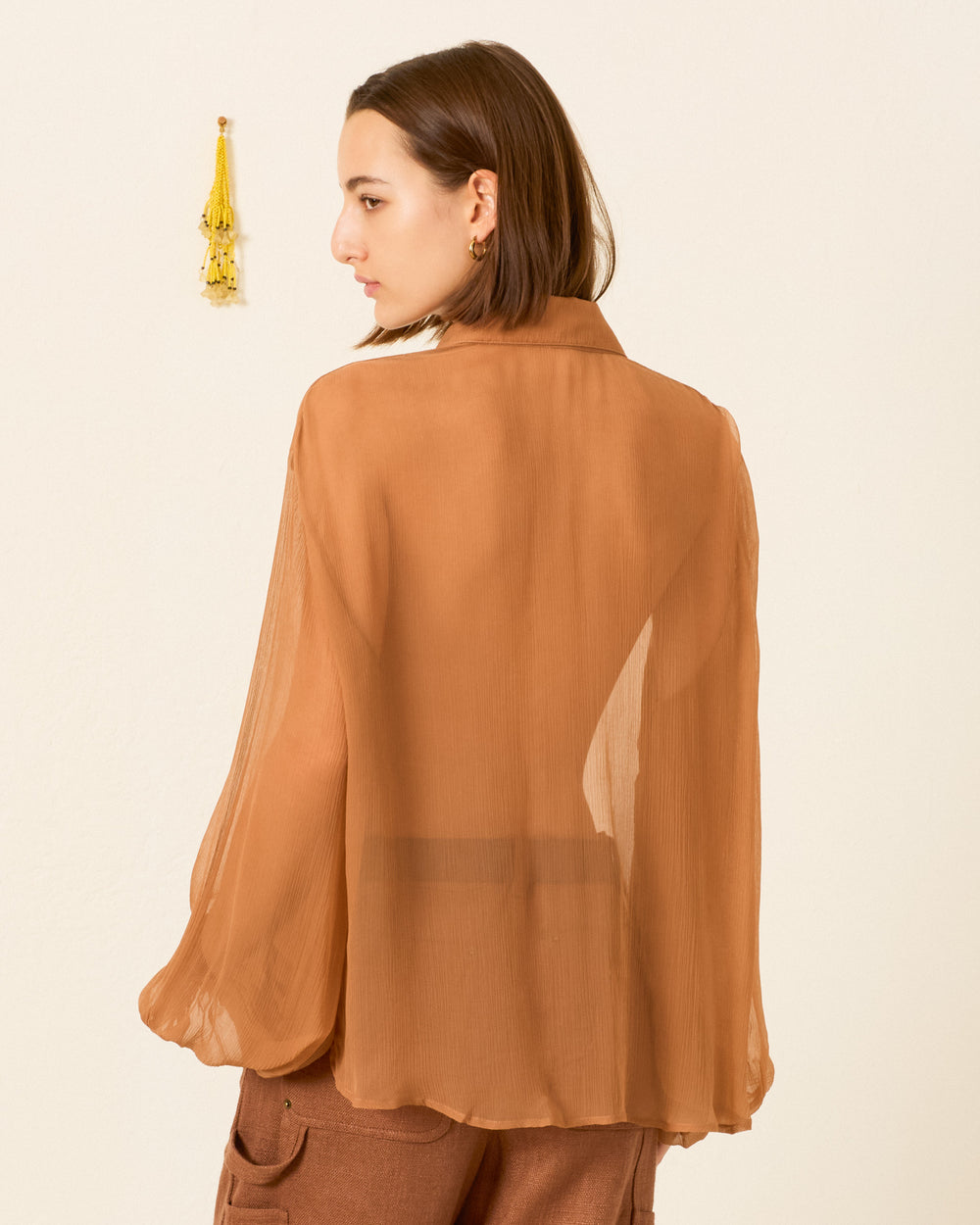 A person with short brown hair wears the Elfie Fawn Chiffon Shirt—a vintage-inspired, sheer, long-sleeve top—and brown pants, standing with their back to the camera near a cream wall with a yellow tassel hanging.