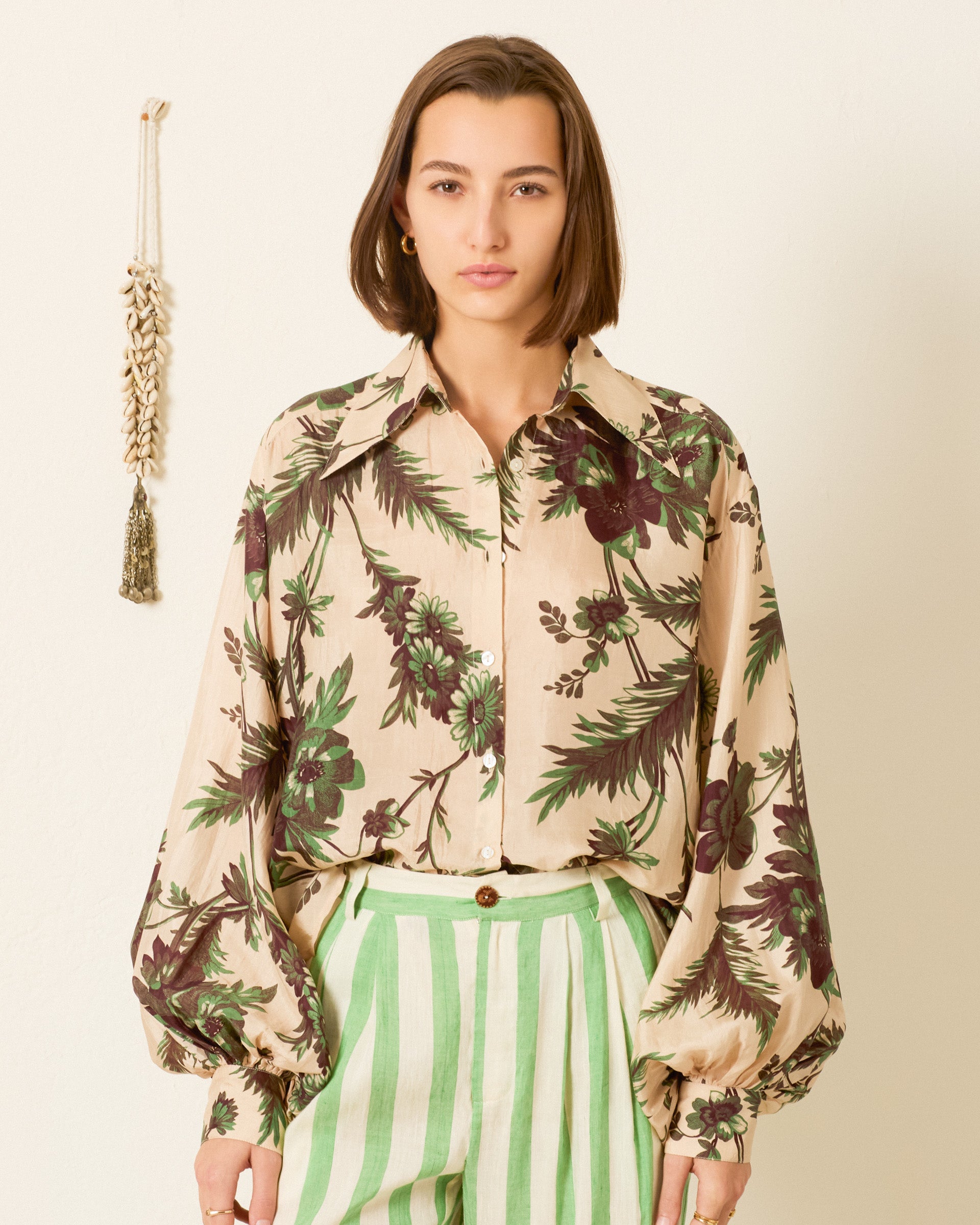 Someone wearing the Elfie Summermoss Silk Shirt with voluminous sleeves and green striped pants stands against a plain background, with a wall decoration visible on the left.