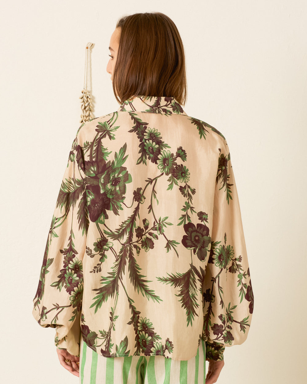 A person faces away, wearing the Elfie Summermoss Silk Shirt featuring a green and brown floral print, paired with green striped pants, against a plain light wall.