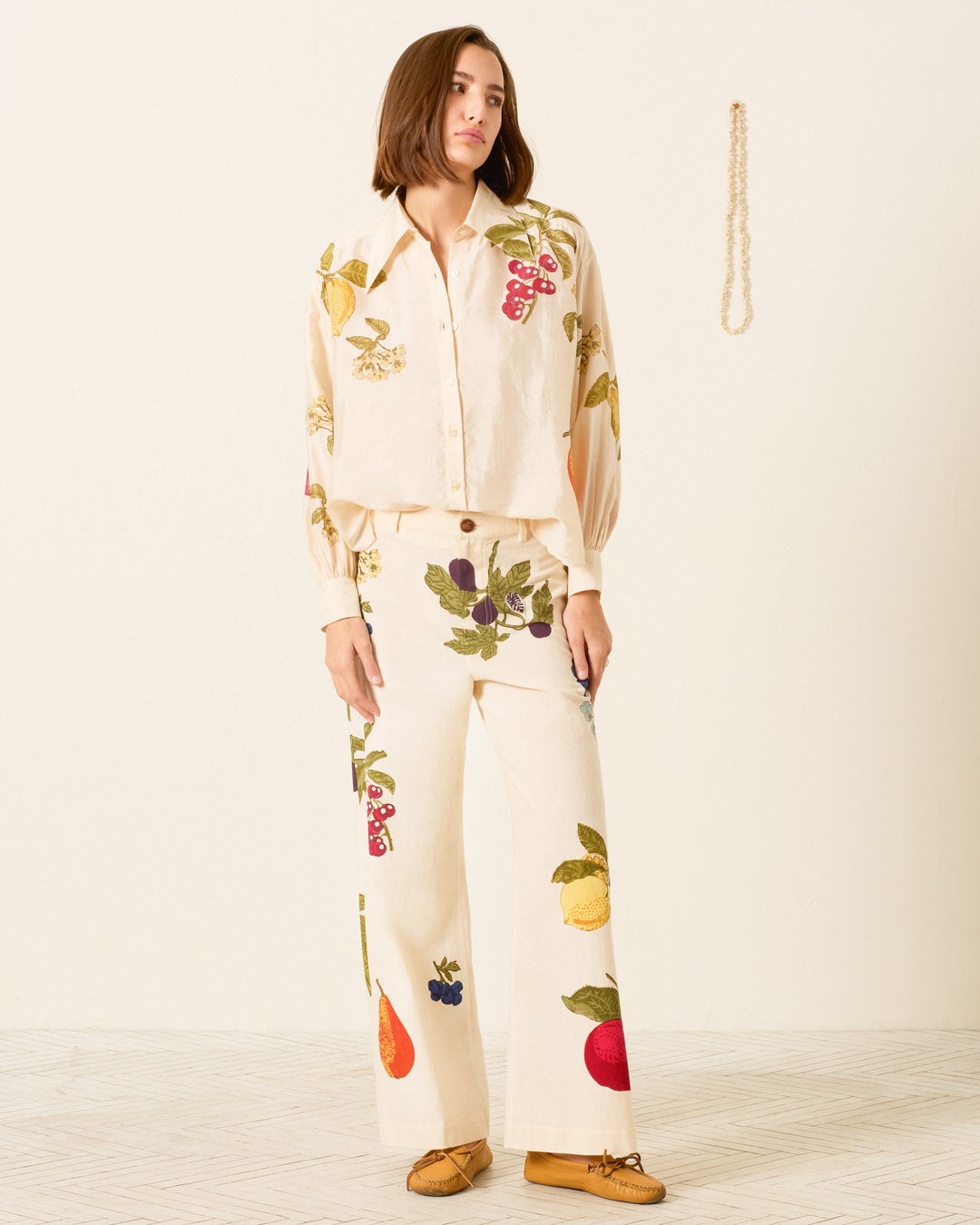 A person stands indoors wearing the Emery Four Fruit Silk Shirt, a cream-colored artisan garment featuring large embroidered fruit designs, paired with tan moccasins.