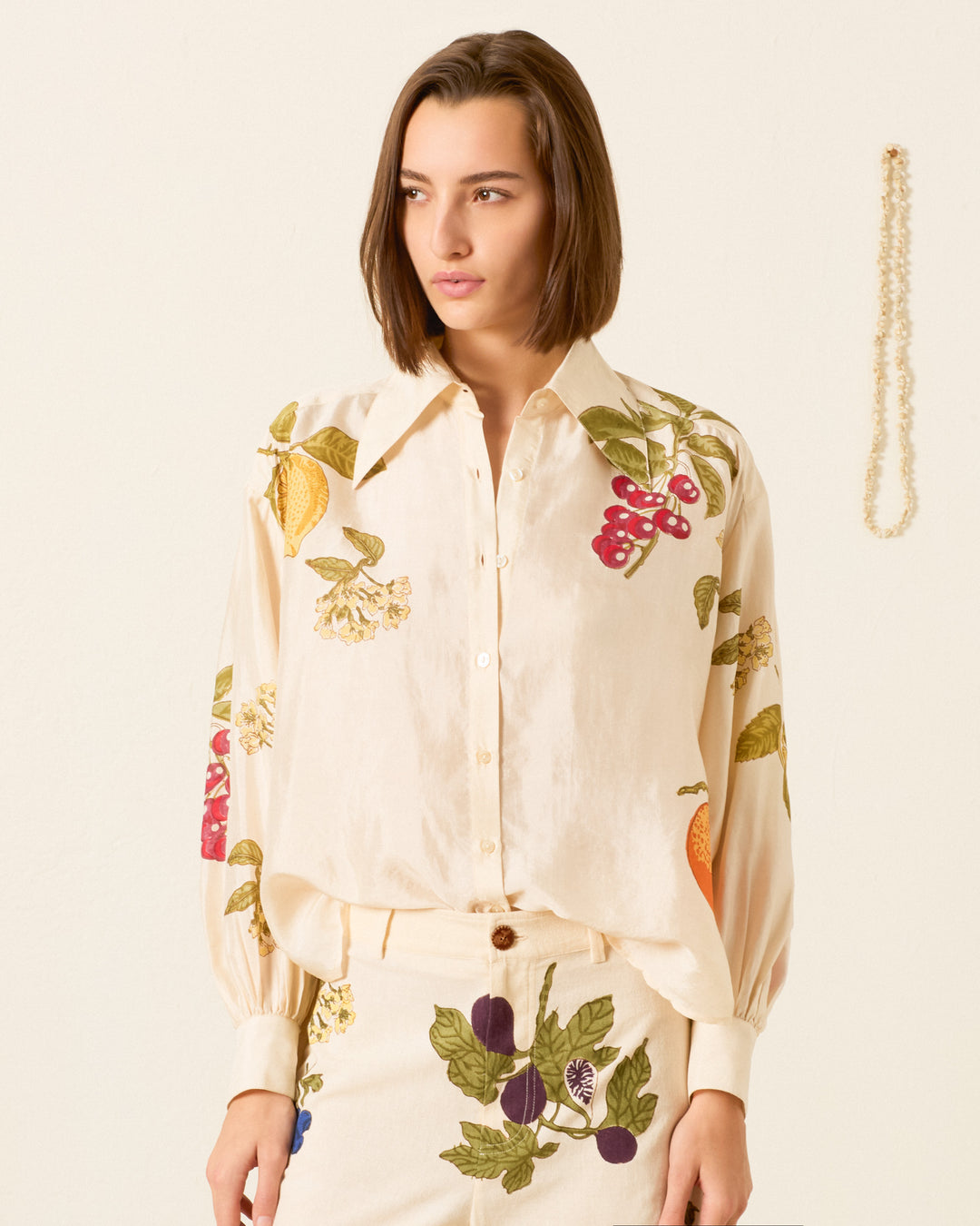 A woman wearing the cream-colored Emery Four Fruit Silk Shirt, featuring colorful fruit prints, stands in front of a plain wall with a pearl necklace hanging on it.